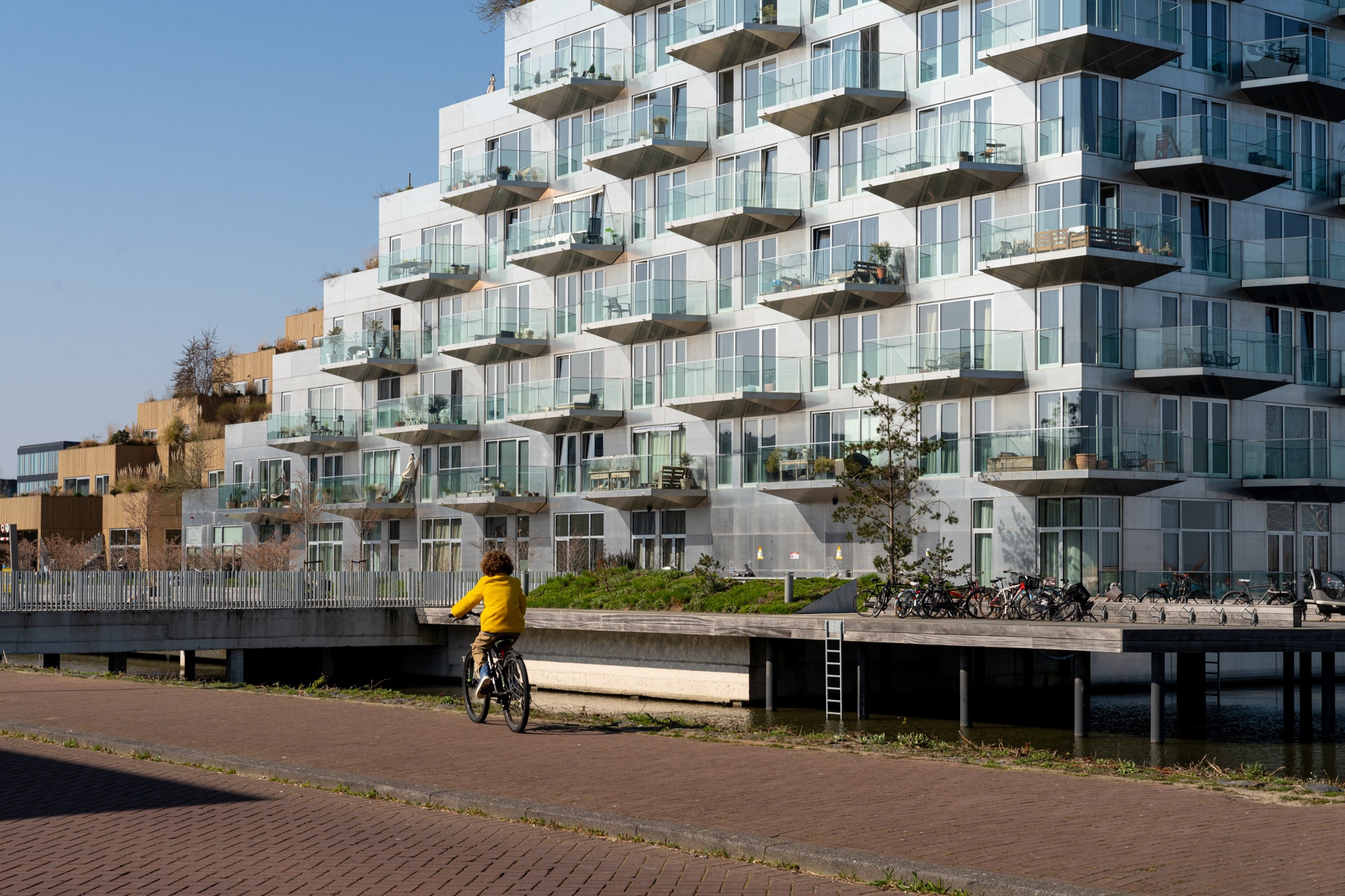 Een persoon met een geel jack rijdt op een fiets langs een modern flatgebouw met talloze glazen balkons op een heldere, zonnige dag.