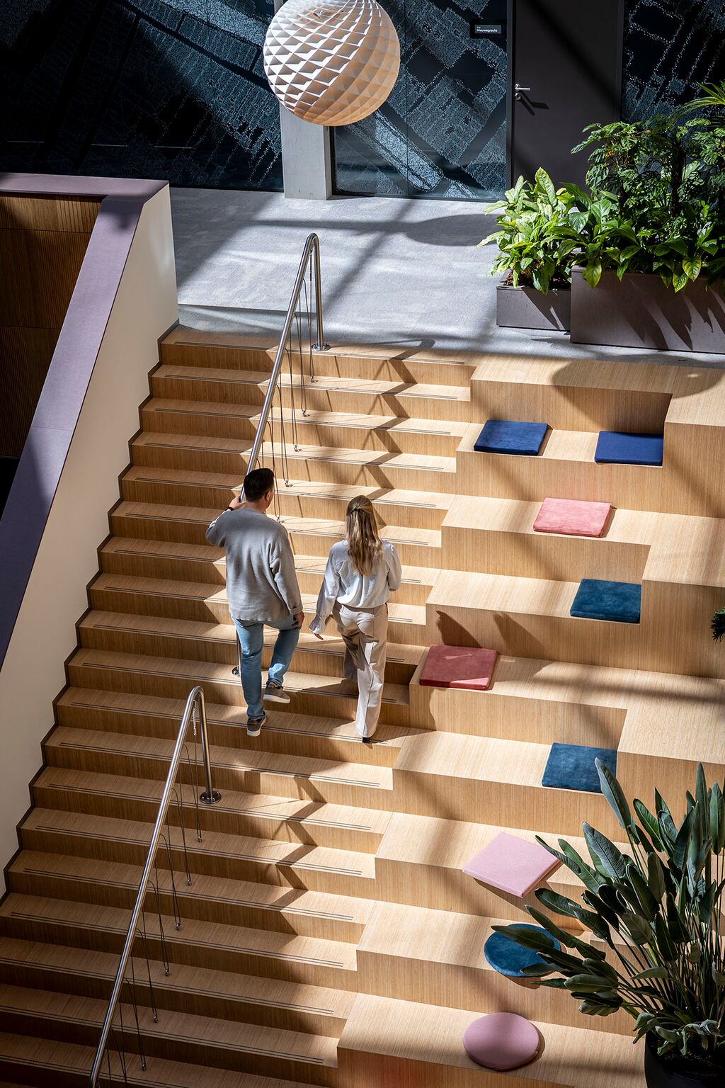 Een man en vrouw lopen een brede houten trap op met kleurrijke vierkante kussens. Zonlicht stroomt naar binnen en verlicht de kamerplanten en moderne inrichting in een ruim, eigentijds interieur.