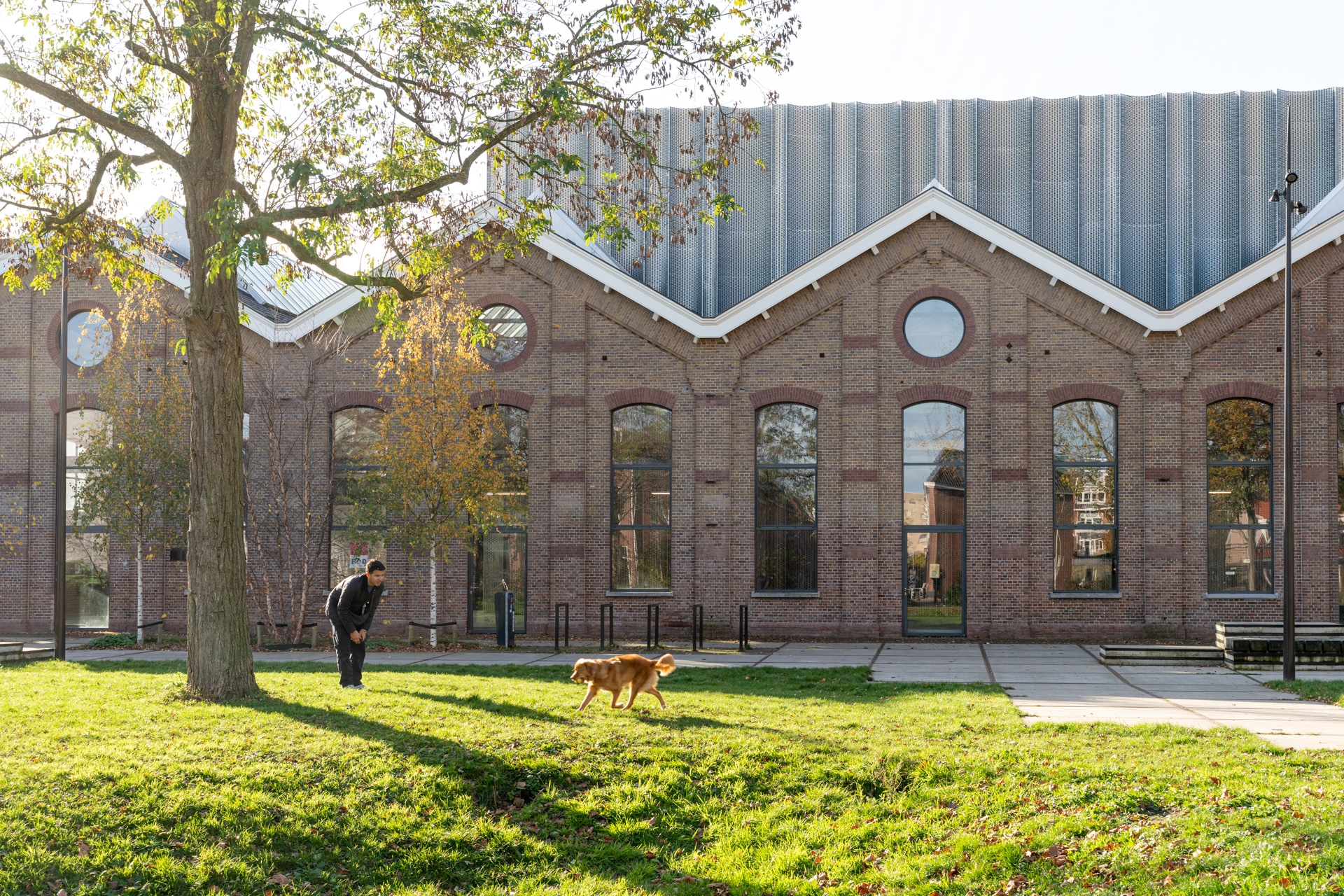 Een persoon en een hond spelen op een grasveld voor een groot bakstenen gebouw met hoge ramen en een metalen dak op een zonnige dag.