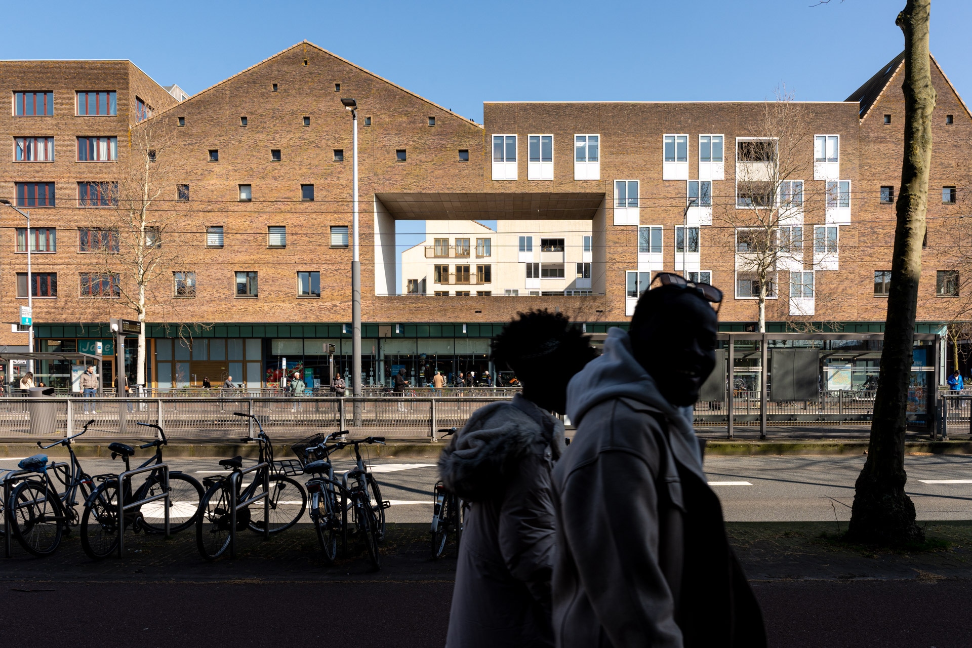 Twee mensen lopen langs geparkeerde fietsen op een stoep voor een modern bakstenen gebouw met grote ramen en geometrische vormen, onder een strakblauwe hemel.