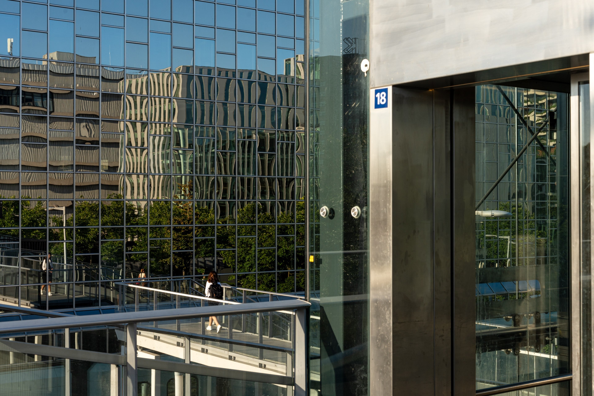 Een moderne stad met reflecterende glazen gebouwen, metalen leuningen en twee mensen die over een skybridge lopen. Zonlicht werpt scherpe schaduwen en reflecties, waardoor een geometrisch patroon op de gebouwen ontstaat.