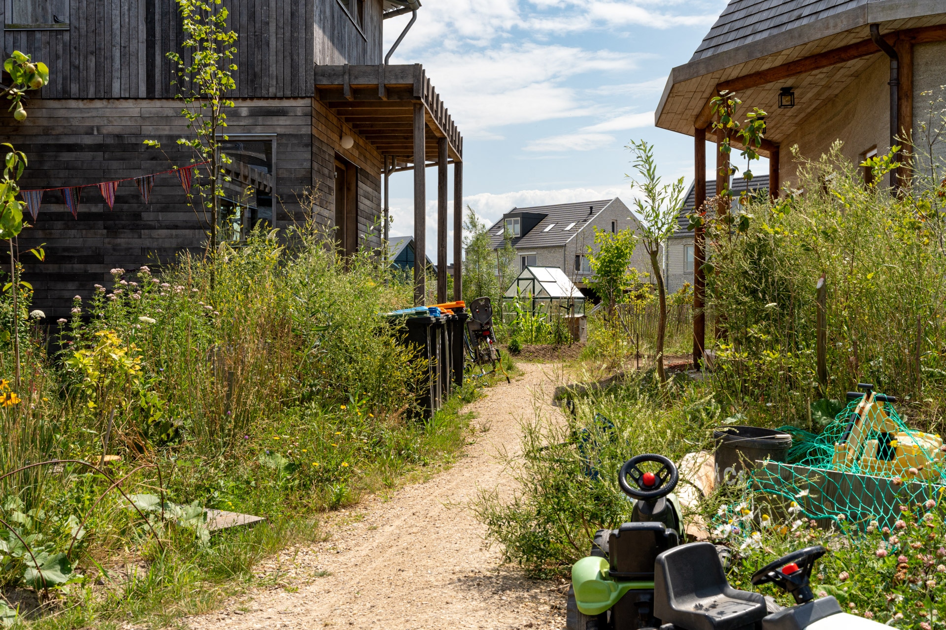 Een smal zandpad loopt tussen twee houten huizen met overwoekerd gras en planten. Verschillende vuilnisbakken staan langs het pad en kinderspeelgoed ligt verspreid op de voorgrond. Op de achtergrond zijn een kas en meer huizen te zien.