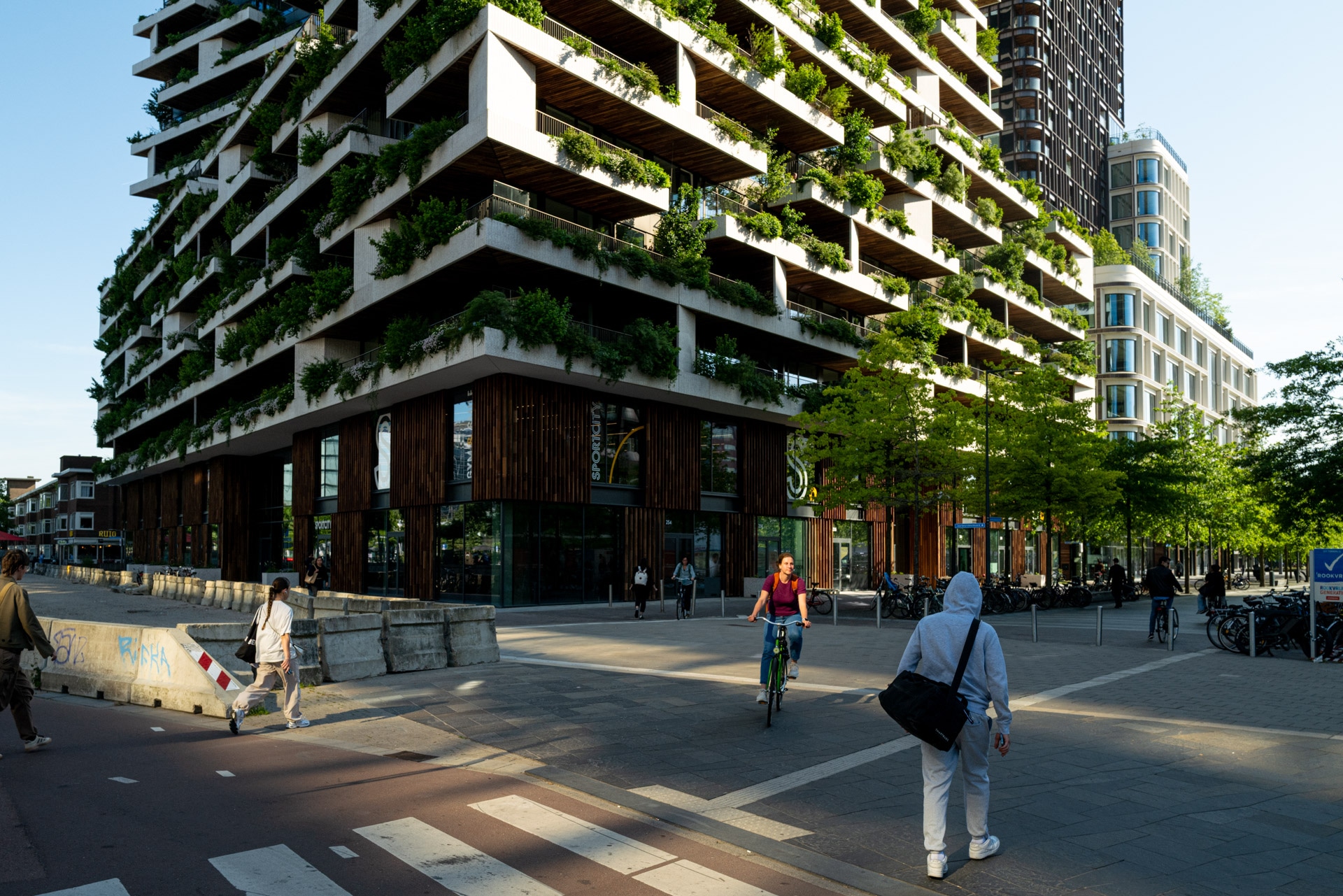 Een modern gebouw met veel balkons bedekt met planten staat op een straathoek; een fietser en verschillende voetgangers steken het zonovergoten kruispunt over omzoomd met bomen en stedelijke infrastructuur.