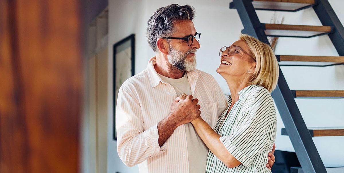 Een ouder koppel lacht gelukkig naar elkaar terwijl ze elkaars hand vasthouden en samen dansen in een gezellig, modern huis met een trap op de achtergrond.
