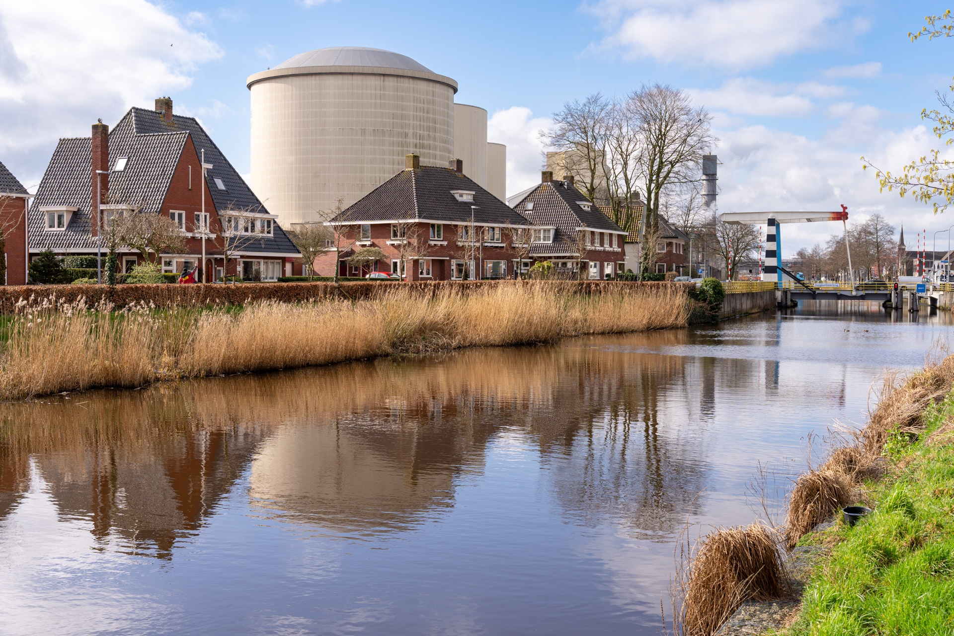 Een kanaal omzoomd door gras en huizen, met een groot rond industrieel gebouw op de achtergrond onder een gedeeltelijk bewolkte hemel, weerspiegeld in het kalme water.