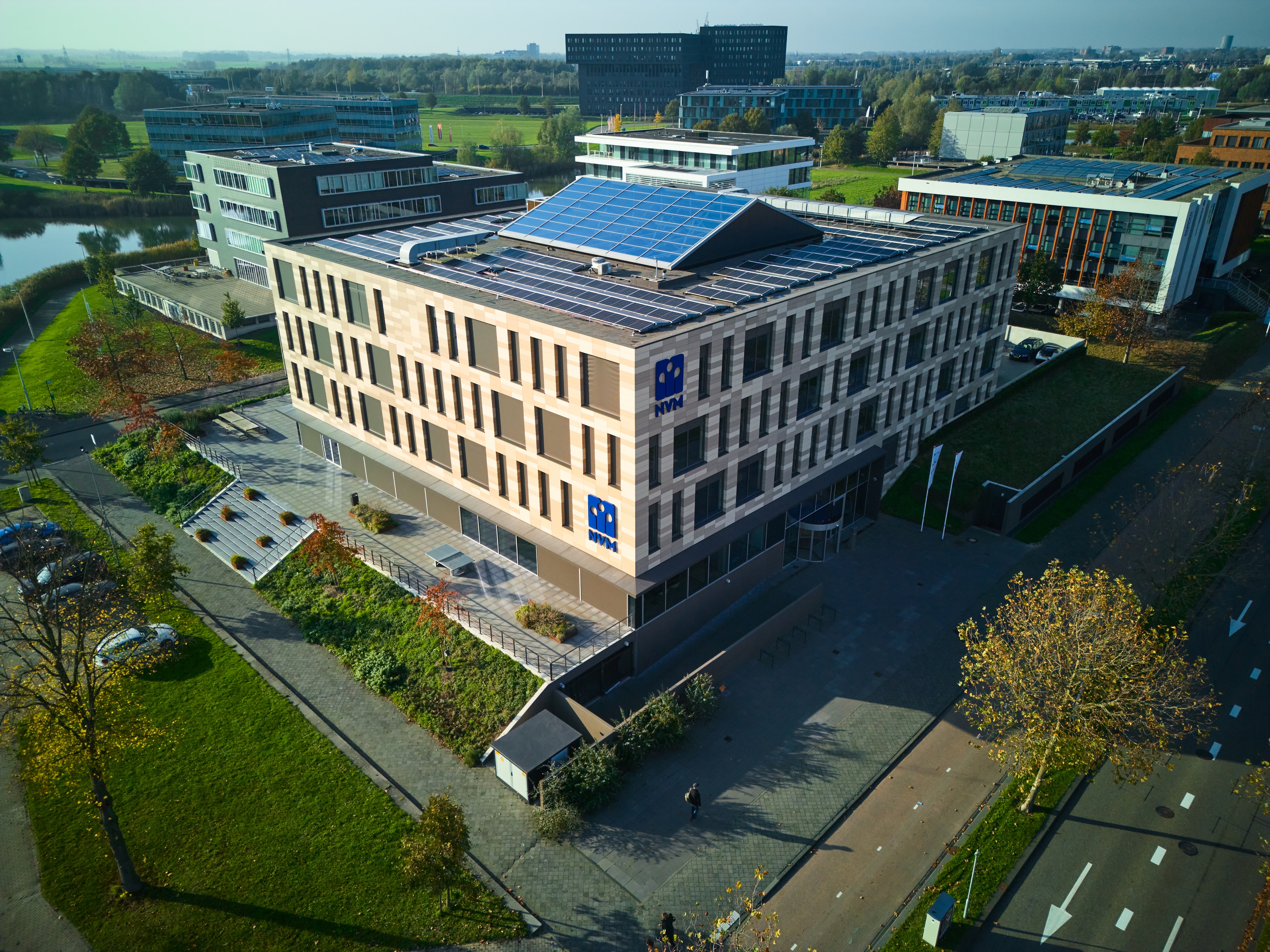 Luchtfoto van een modern kantoorgebouw met meerdere verdiepingen en zonnepanelen op het dak, omgeven door groen, wegen en andere kantoorgebouwen onder een heldere hemel.