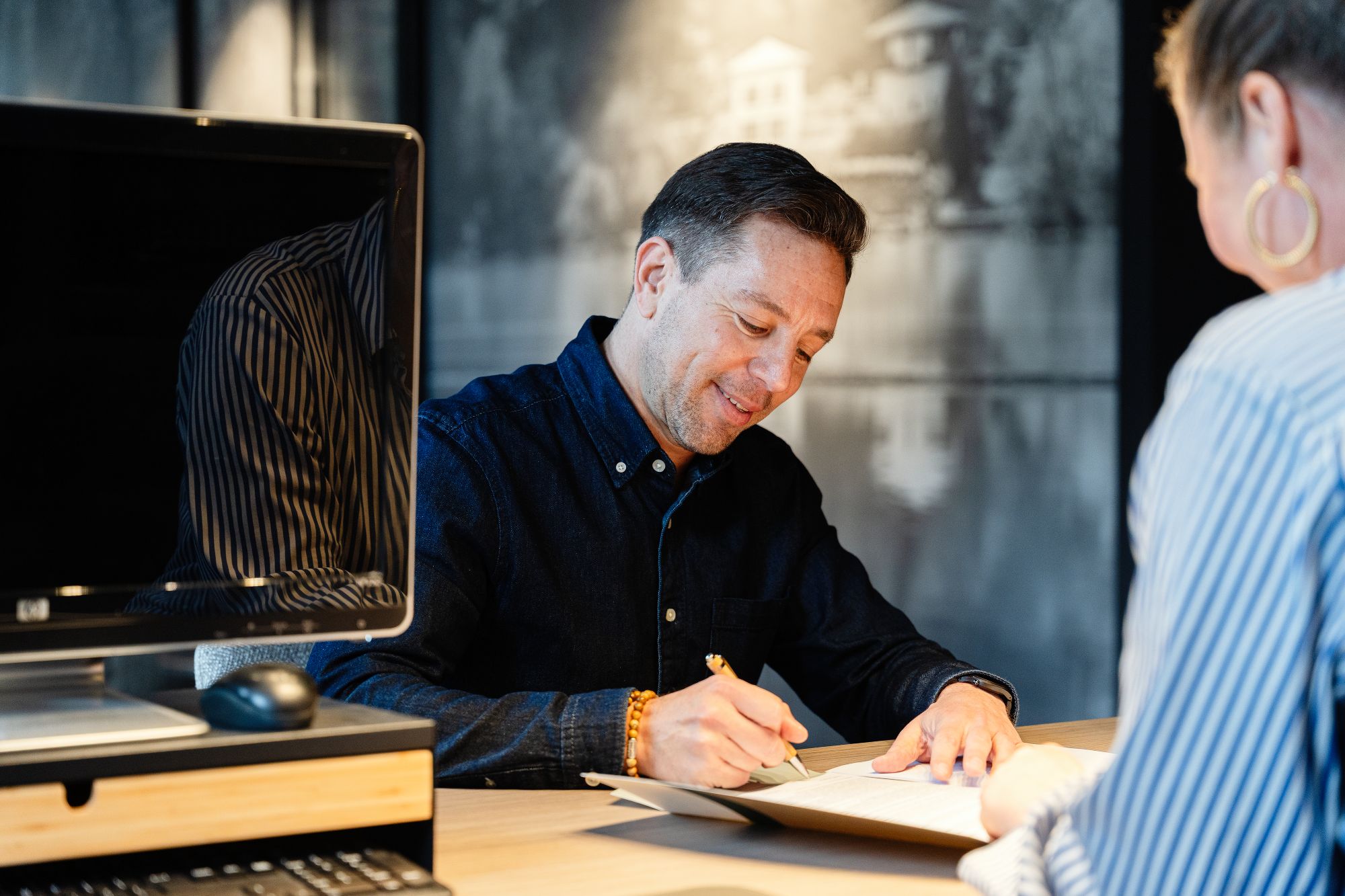 Een man zit aan een bureau, glimlacht terwijl hij een document ondertekent, met een andere persoon tegenover hem onscherp; een computermonitor staat op het bureau naast hem.