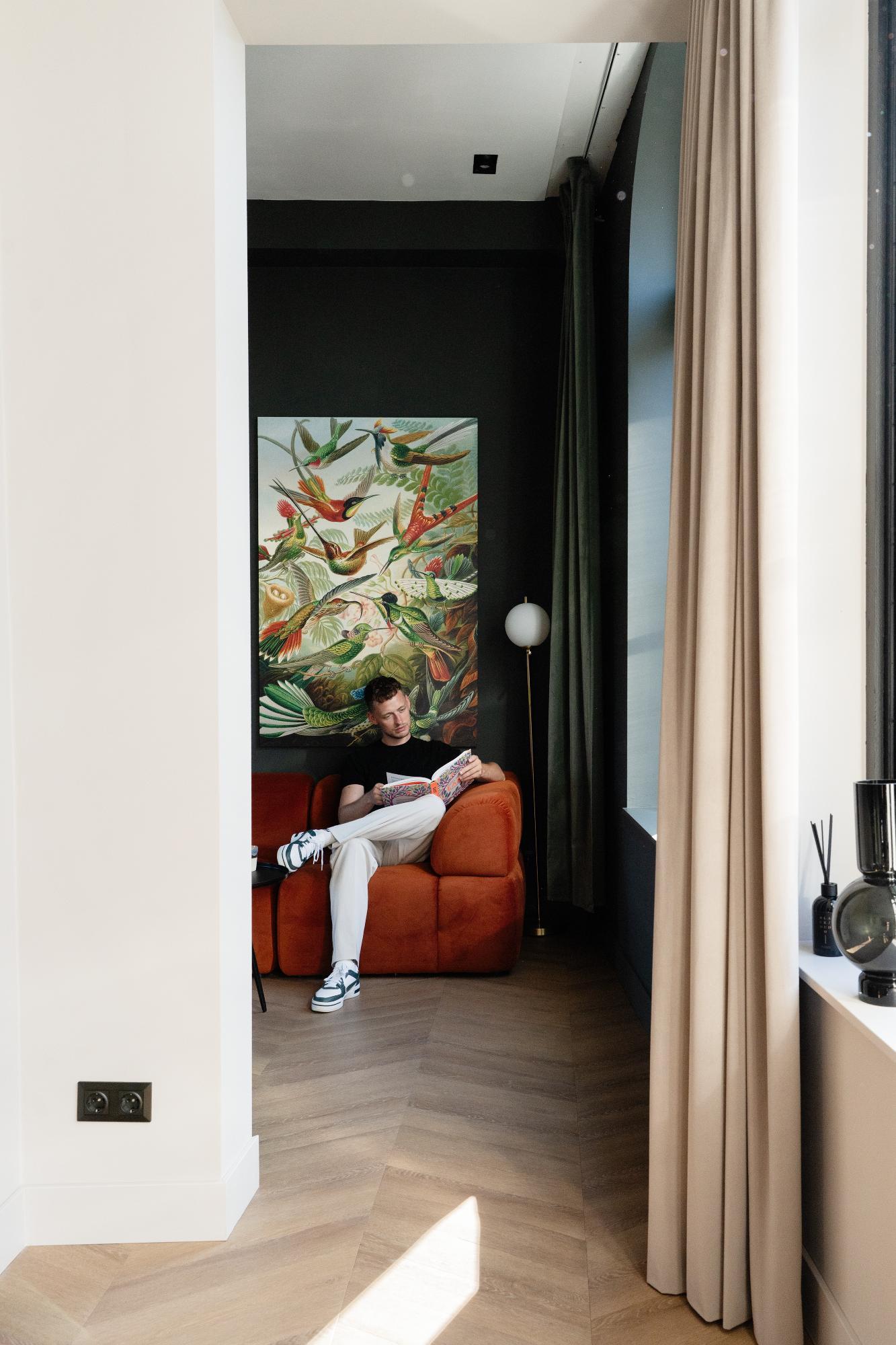 Een man zit te lezen op een oranje sofa in een moderne kamer met houten vloeren, grote ramen, beige gordijnen en een kleurrijk schilderij van vogels op de donkere muur achter hem.