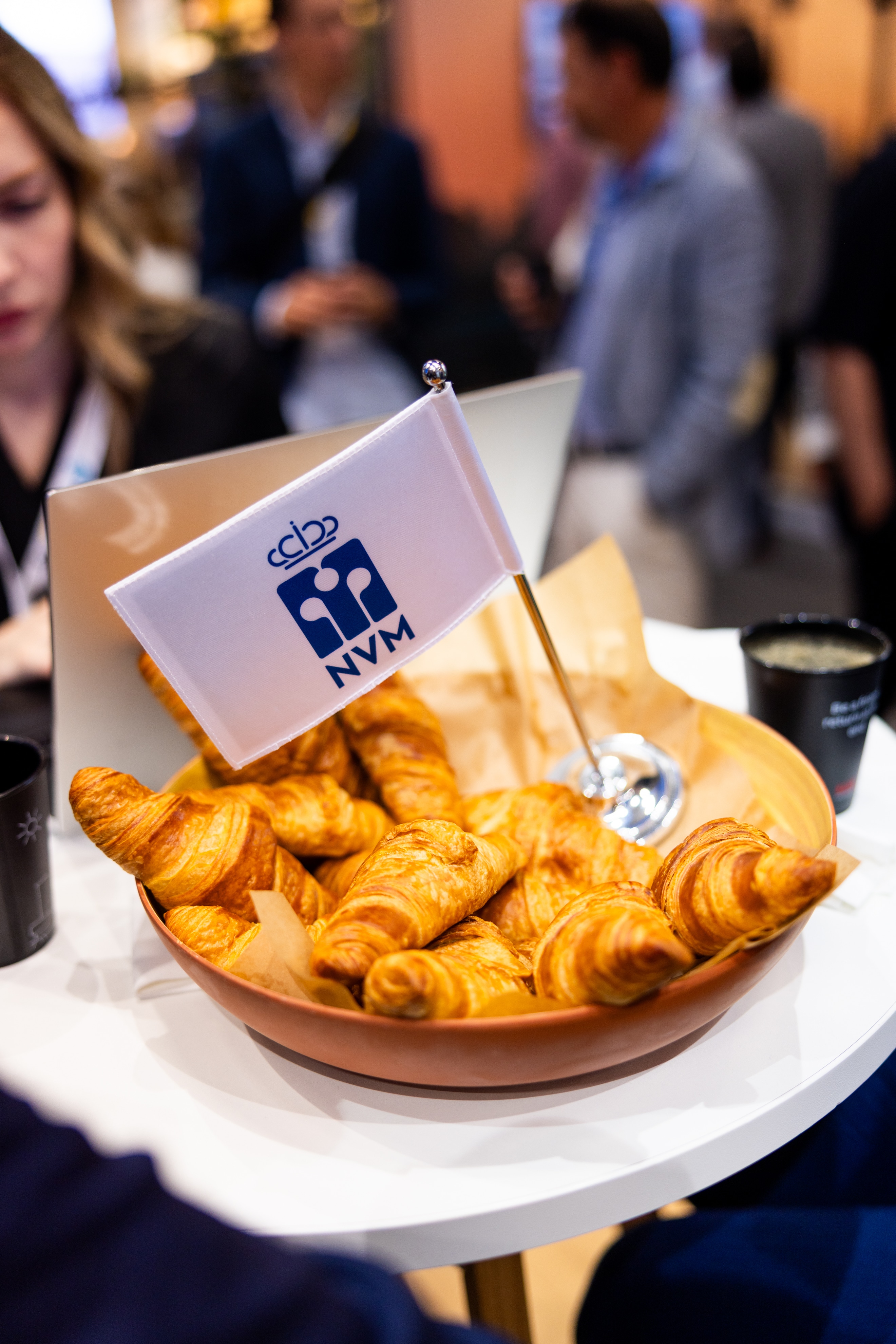 Een bord croissants met een NVM-vlaggetje op een tafel tijdens een evenement, omringd door mensen in zakelijke kleding en koffiekopjes.