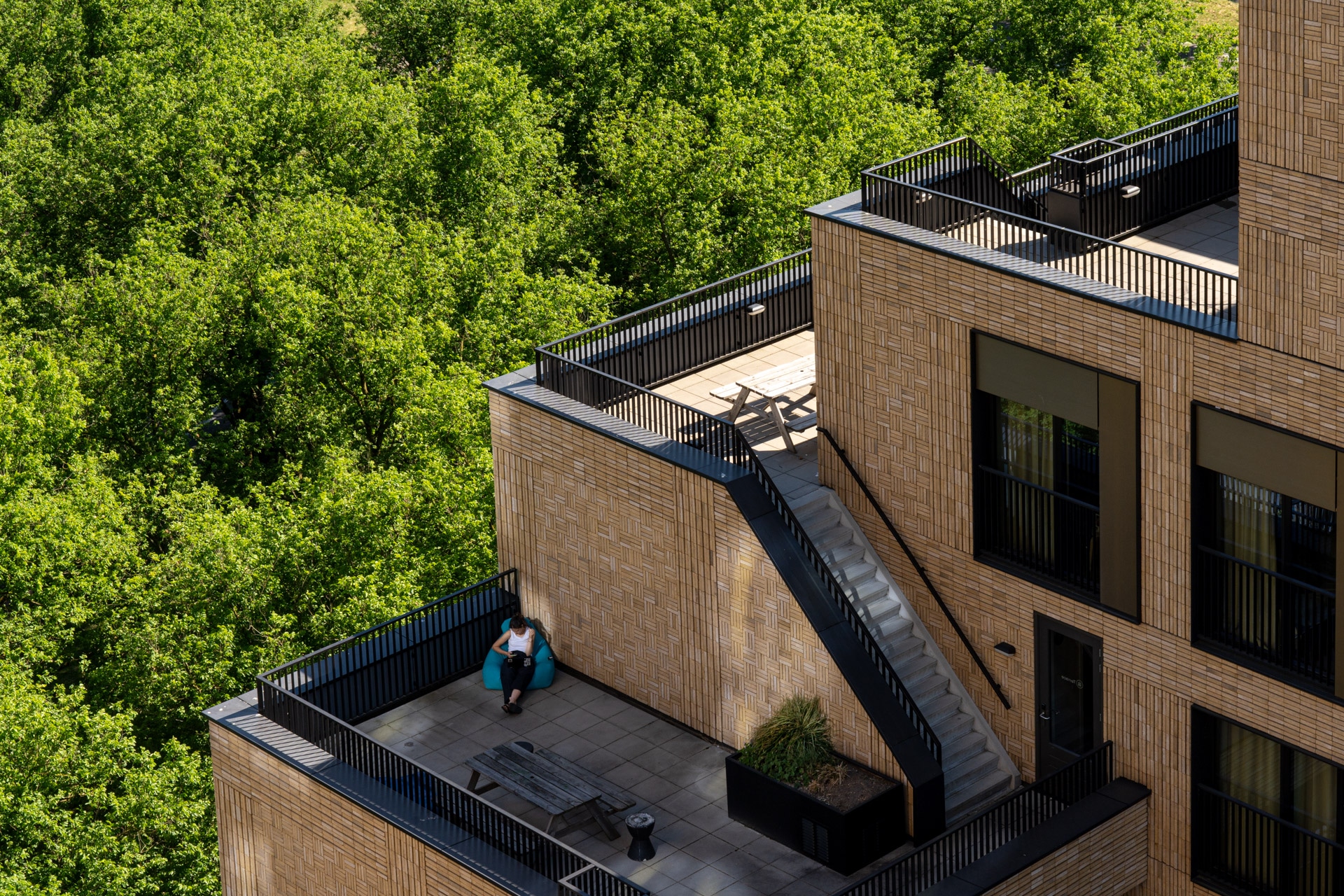 Een persoon zit alleen op een stoel op een dakterras van een modern bakstenen gebouw, omringd door zwarte balustrades, met weelderig groene bomen op de achtergrond.