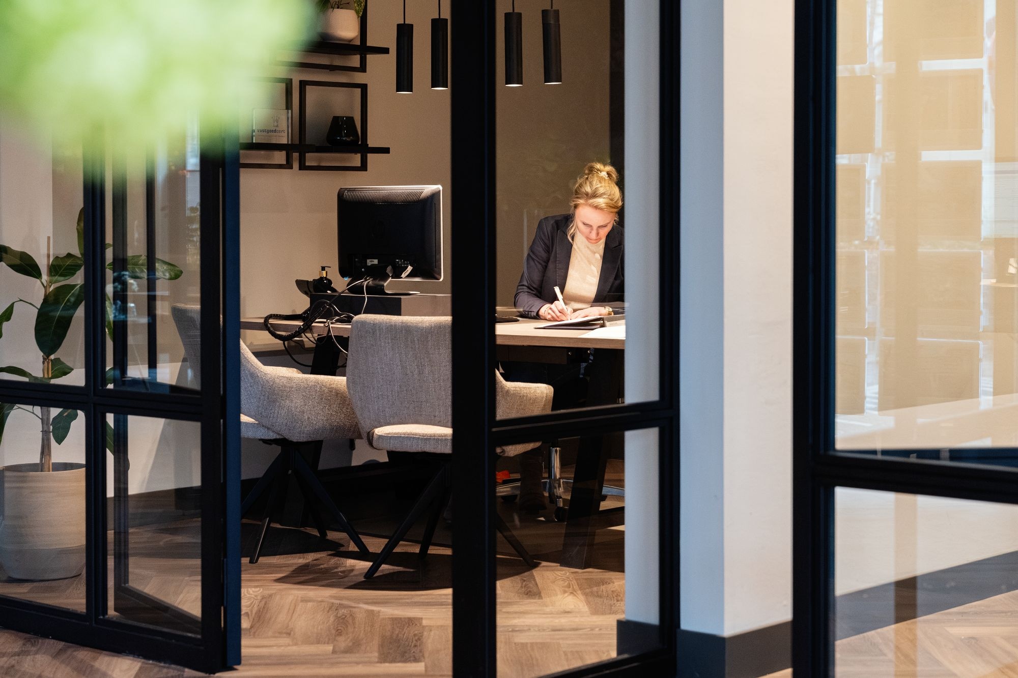 Een vrouw zit alleen achter een bureau in een modern kantoor en schrijft in een notitieblok. Het kantoor heeft grote glazen muren, een computerscherm, grijze stoelen en een moderne inrichting met planten en donkere verlichting.