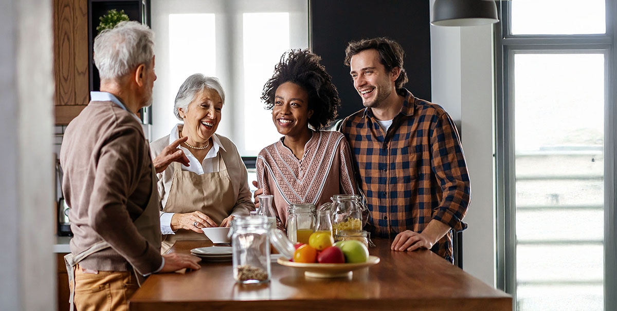 Vier mensen staan samen in een lichte keuken, lachend en pratend rond een aanrecht met fruit en potjes. Twee oudere volwassenen en twee jongere volwassenen lijken van elkaars gezelschap te genieten in een ontspannen, vriendelijke sfeer.