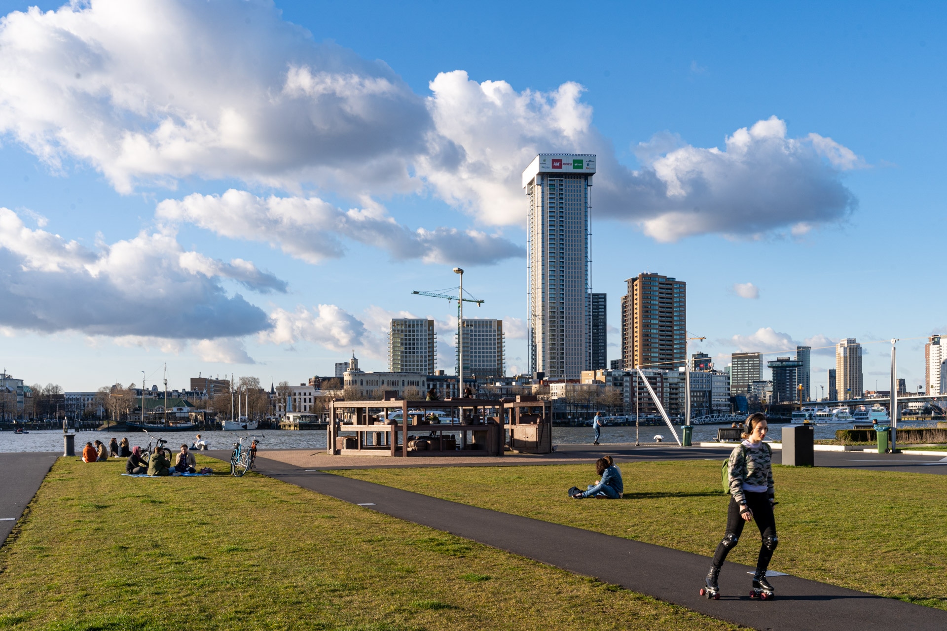 Mensen ontspannen op gras bij een pad langs de rivier onder een blauwe lucht met wolken; een persoon rolschaatst op de voorgrond met hoge stadsgebouwen en een kraan zichtbaar op de achtergrond.