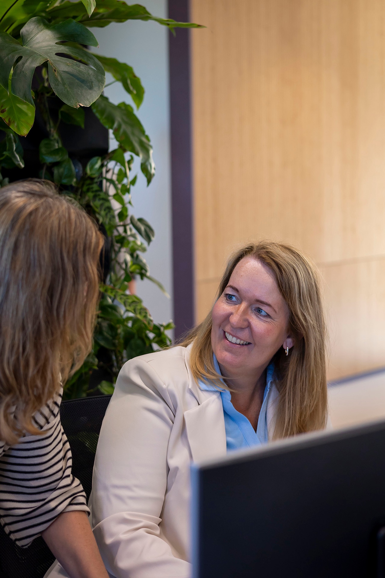 Twee vrouwen zitten te praten in een kantooromgeving. De ene draagt een witte blazer en glimlacht hartelijk naar de andere, die gekleed is in een gestreept overhemd. Op de achtergrond zijn groene planten en een houten muur te zien.