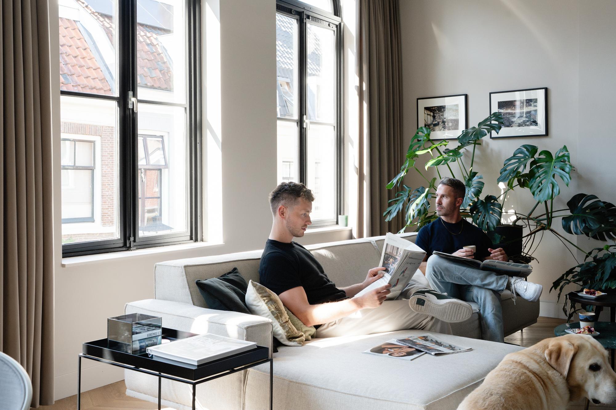 Twee mannen ontspannen zich in een lichte woonkamer met grote ramen; de een leest een krant op een bank terwijl de ander met een laptop zit. Een hond staat op de voorgrond en bladplanten en ingelijste foto's sieren de ruimte.