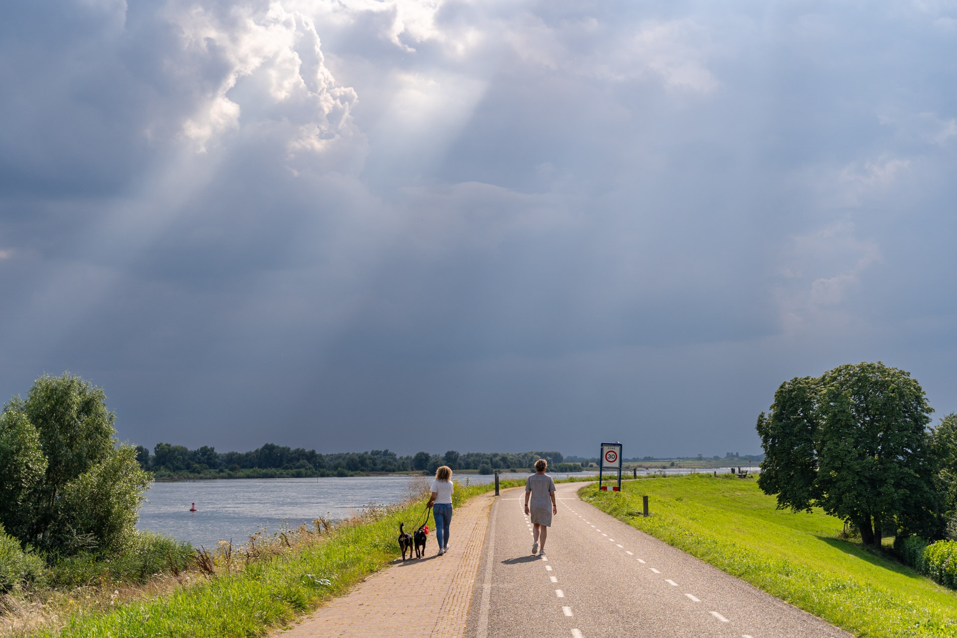 Twee mensen lopen langs een pad langs de rivier onder een bewolkte hemel waar zonnestralen doorheen schijnen. De ene persoon loopt met honden, terwijl de andere vooruit loopt. Bomen en groen gras omzomen het pad en links is de rivier zichtbaar.