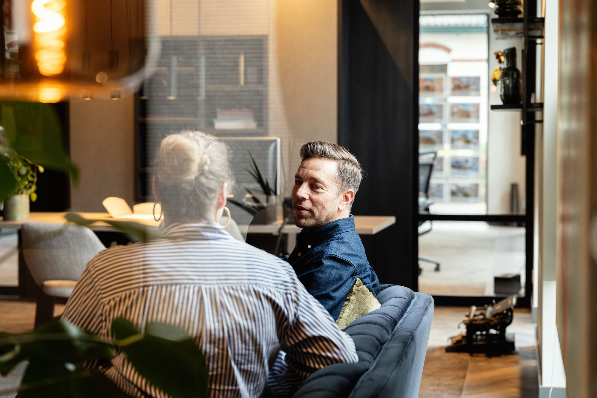 Een man en een vrouw zitten op een blauwe bank in een modern, goed verlicht kantoor en zijn in gesprek. Het kantoor heeft glazen muren, planken en een bureau op de achtergrond.