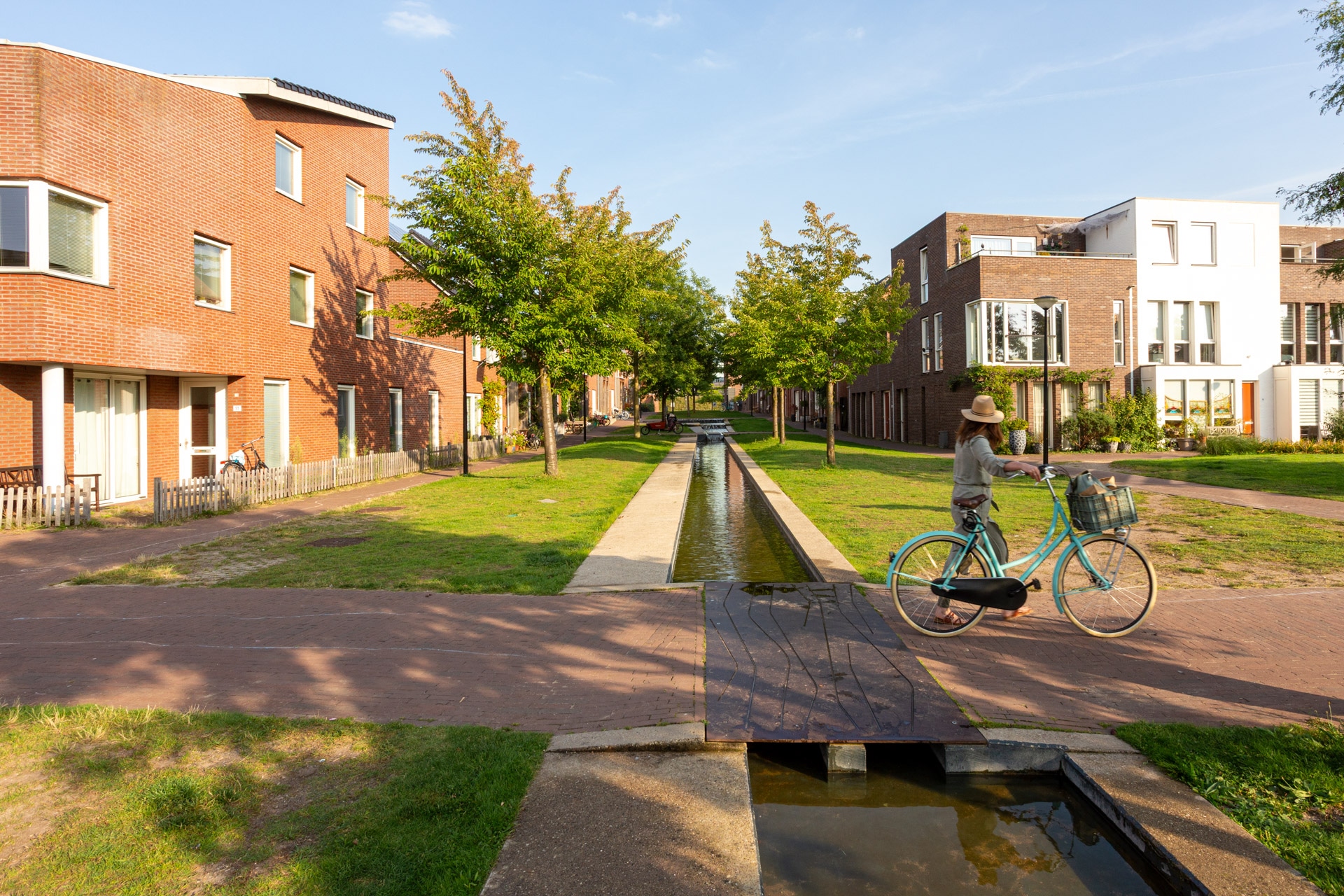 Een persoon met een hoed rijdt op een fiets over een pad langs een smalle gracht in een woonwijk met moderne bakstenen gebouwen, bomen en groen gras op een zonnige dag.