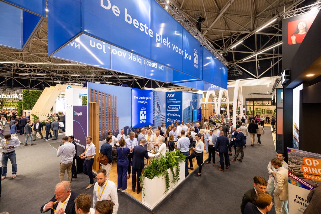 Een drukke beursstand met veel netwerkende en pratende mensen. Boven de stand hangen grote blauwe borden met Nederlandse tekst. De stand is voorzien van planten, hoge displaypanelen en merkbanners. De locatie heeft hoge plafonds en heldere verlichting.