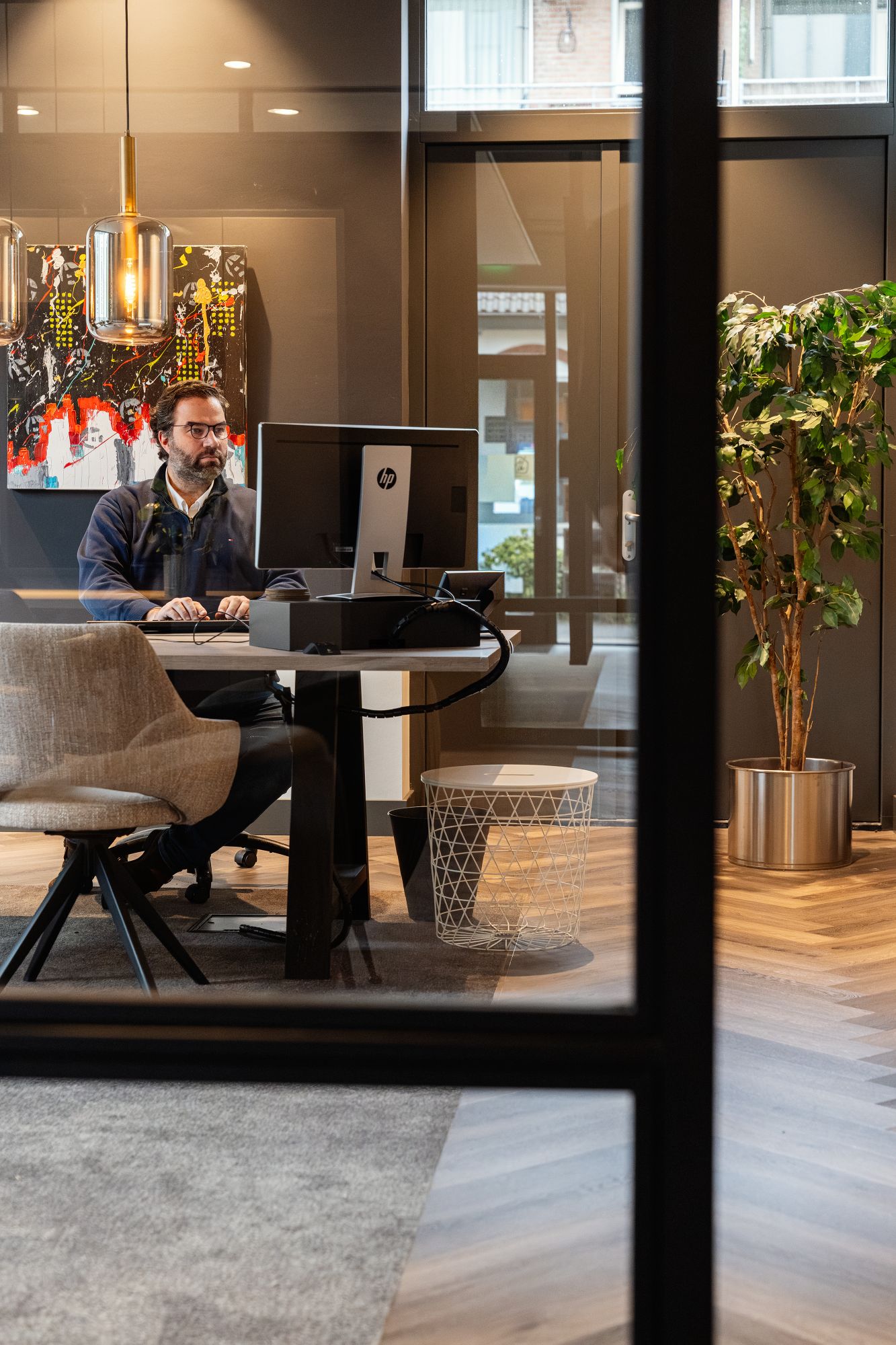 Een man met bril zit aan een modern bureau te werken aan een desktopcomputer in een stijlvol kantoor met een grote plant, abstracte muurkunst, hanglampen en houten vloeren.