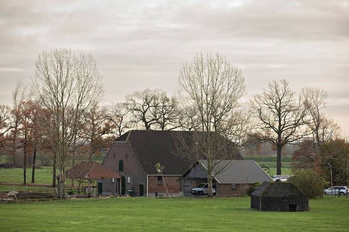 Een rustig landelijk tafereel met een bakstenen boerderij, een paar bijgebouwen, bladerloze bomen en een groen grasveld onder een bewolkte hemel. Een auto staat geparkeerd bij het huis en hekken omlijnen delen van het terrein.