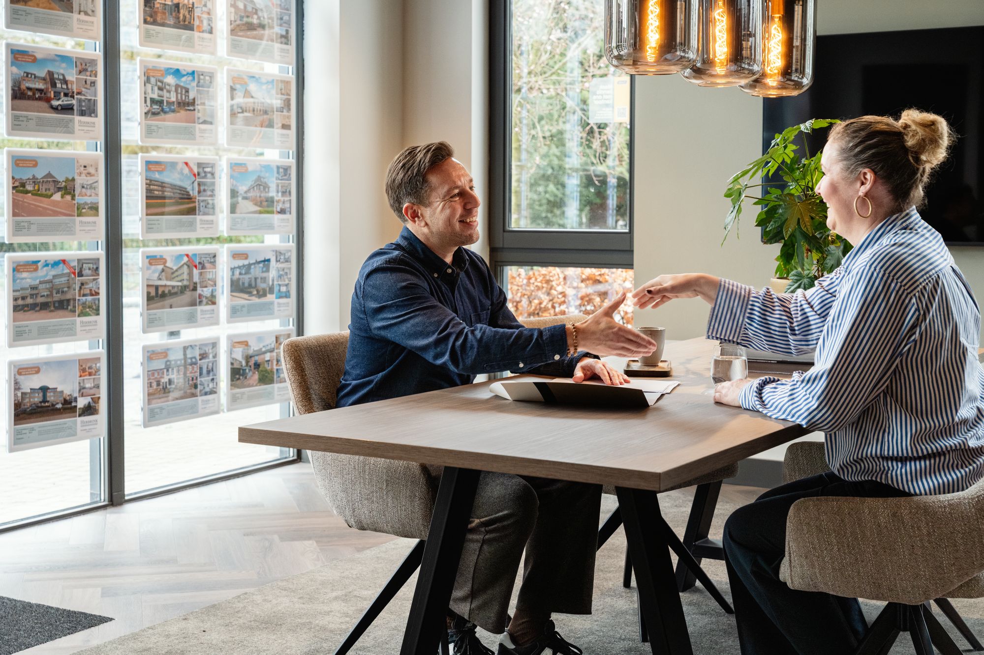Een man en een vrouw zitten tegenover elkaar aan een bureau, schudden elkaar de hand en glimlachen. Ze lijken zich in een modern kantoor te bevinden met grote ramen en onroerend goed advertenties aan de muur.
