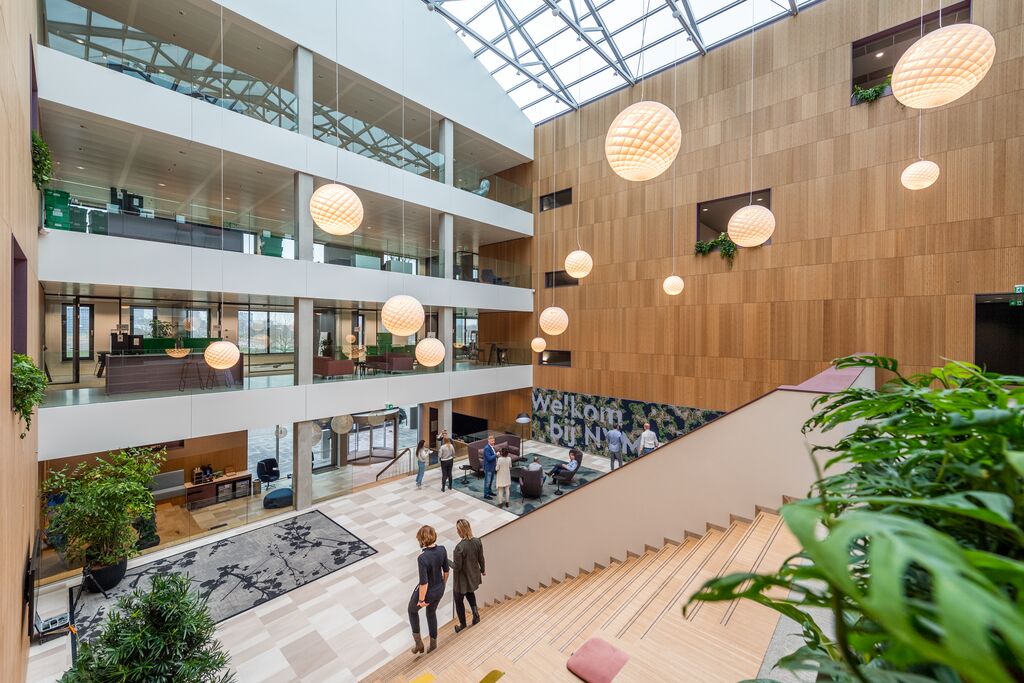 Modern, ruim atrium met houten muren, grote globe hanglampen, kamerplanten en ramen van vloer tot plafond. Verschillende mensen zijn in gesprek in de buurt van de lobby en het trappenhuis. Het bord Welkom bij NN hangt aan de muur.