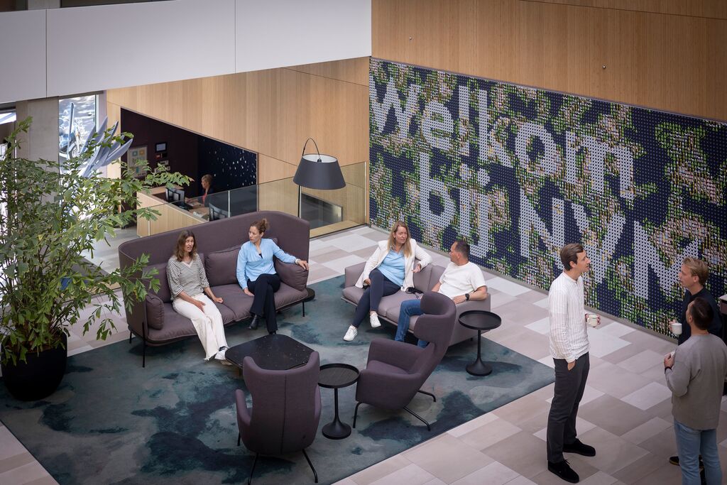 Een groep mensen maakt kennis met elkaar in een moderne loungeruimte met banken en stoelen, een grote muurschildering met de tekst Welkom bij NN en een hoge kamerplant. De ruimte is licht met licht hout en witte muren.