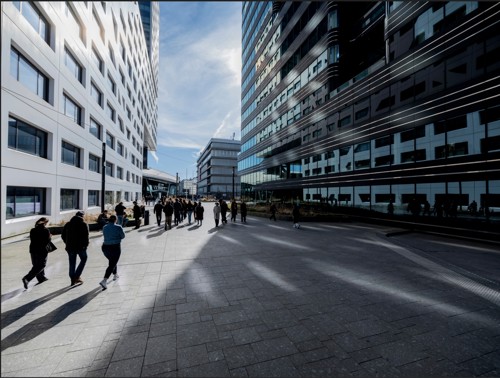 Een groep mensen loopt tussen moderne kantoorgebouwen op een zonnige dag, lange schaduwen werpend op de brede, geplaveide stoep. De lucht is blauw met wat lichte wolken.