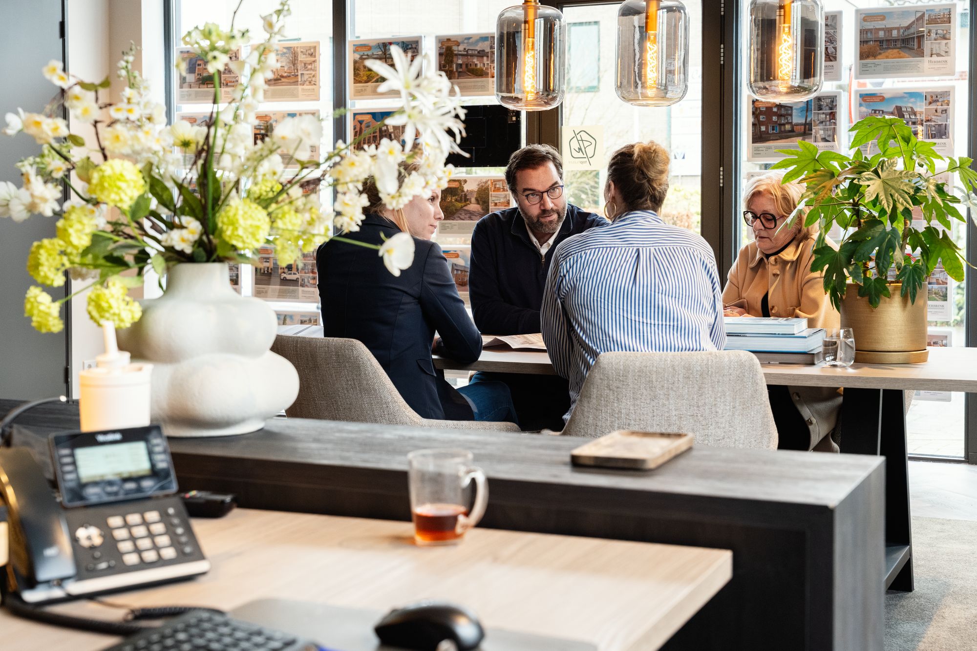 Vier mensen zitten rond een tafel in een modern kantoor en zijn in gesprek. De werkruimte heeft grote ramen, planten, bloemen en een bureau met een telefoon en een mok op de voorgrond.