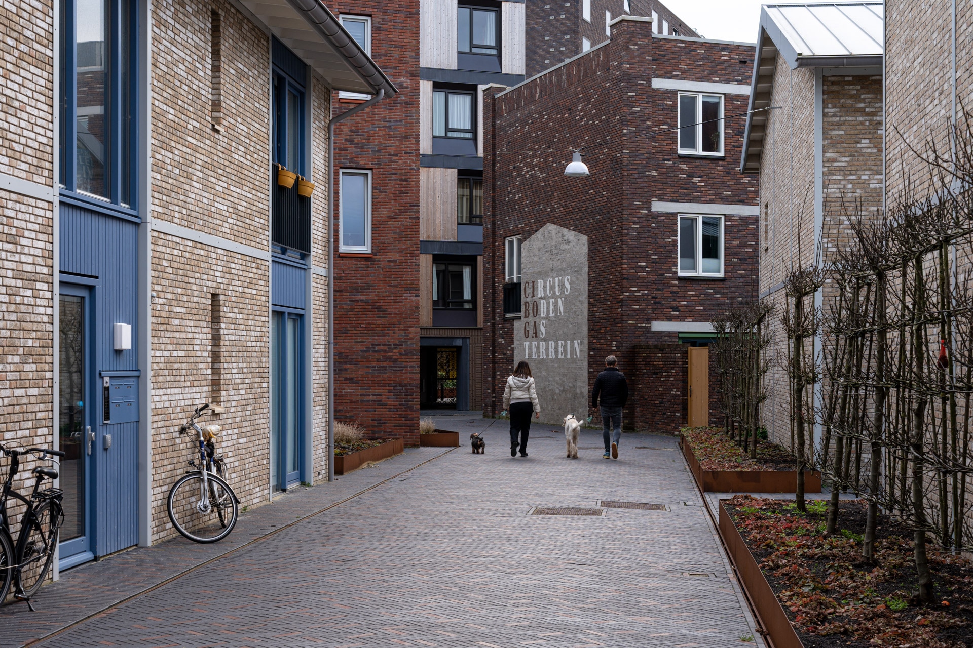 Een smalle, met bakstenen geplaveide woonstraat met moderne bakstenen gebouwen. Twee mensen lopen met honden in de richting van een muur met Nederlands schrift. Links staan twee fietsen geparkeerd en rechts groeien kale planten.