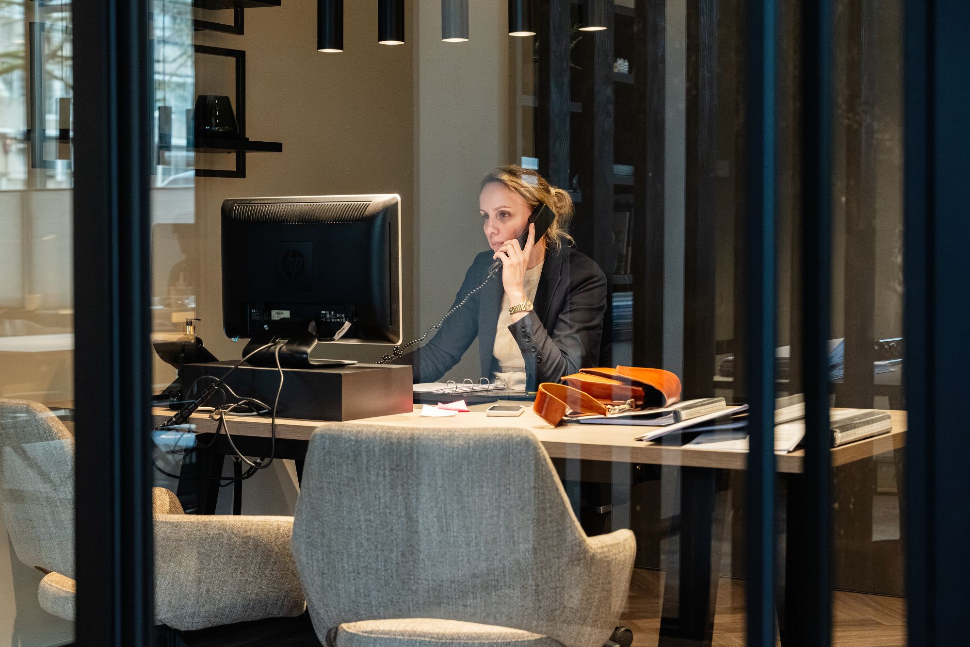 Een vrouw zit aan een bureau in een modern kantoor, telefoneert terwijl ze naar een computerscherm kijkt. Op het bureau liggen papieren, een notitieblok en een tas. De scène wordt bekeken door glazen wanden.