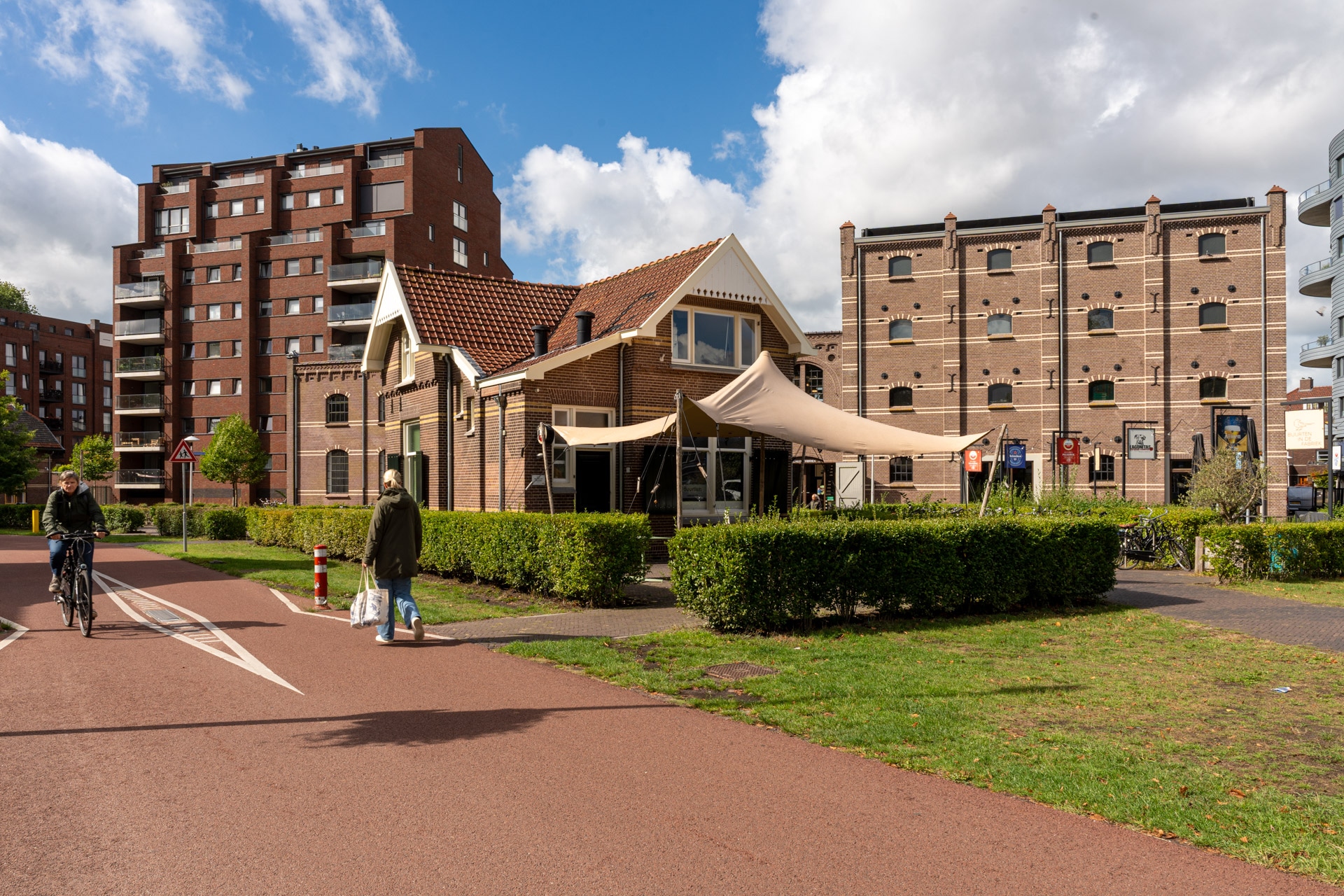 Een klein bakstenen café met een beige luifel staat aan een rood fietspad, omringd door heggen. Moderne en historische flatgebouwen staan op de achtergrond. Mensen wandelen en fietsen onder een gedeeltelijk bewolkte hemel.