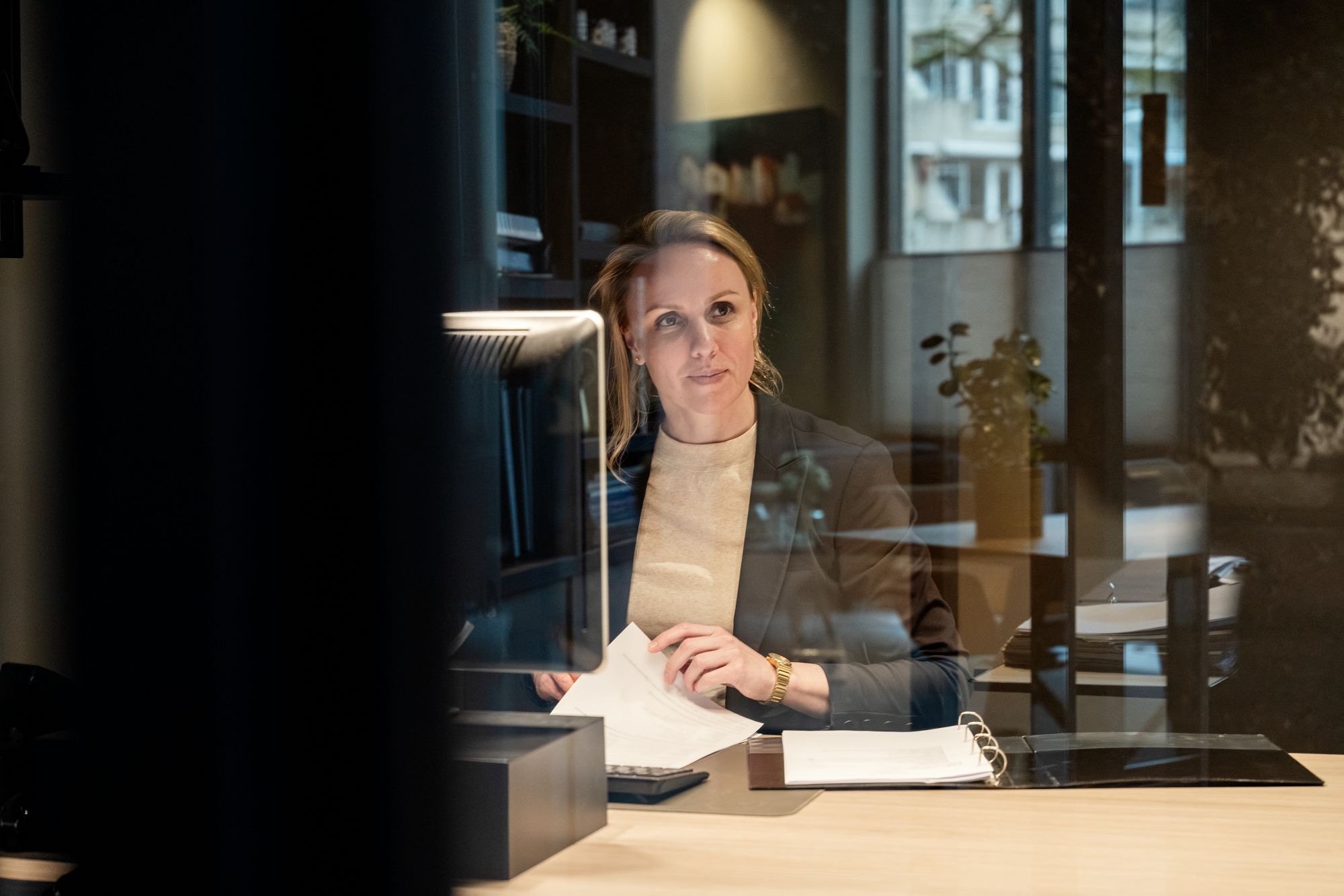 Een vrouw in zakelijke kleding zit aan een bureau in een modern kantoor, houdt papieren vast en kijkt nadenkend naar een computerscherm, waarop weerspiegelingen van ramen en stadsgebouwen zichtbaar zijn.