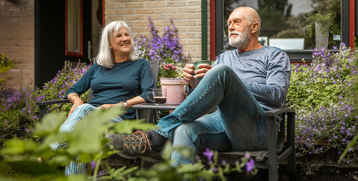 Een oudere vrouw en man zitten samen op een bankje in de tuin, glimlachen en genieten van een drankje. Paarse bloemen en groen omringen hen en op de achtergrond staat een huis met een raam.