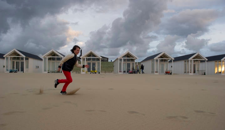 Een persoon met een zwart-witte jas en een rode broek rent over een zandstrand voor een aantal witte strandcabines onder een bewolkte hemel.