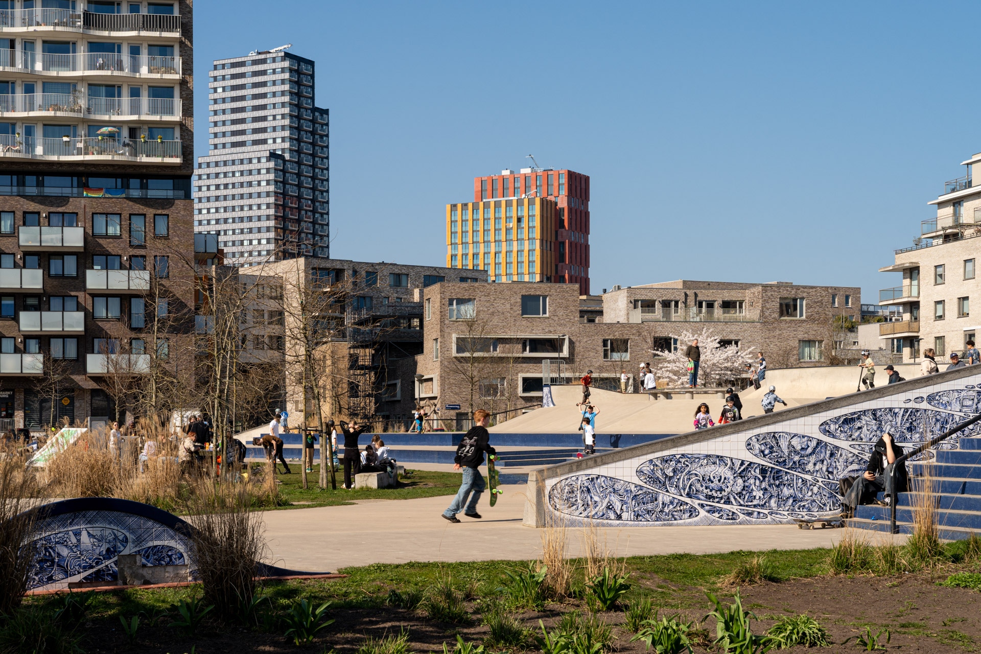 Mensen genieten van een zonnige dag in een stadspark met moderne flatgebouwen op de achtergrond. Sommigen skateboarden op ramps versierd met blauwe en witte tegelpatronen, terwijl anderen ontspannen of rondwandelen in de groene omgeving.