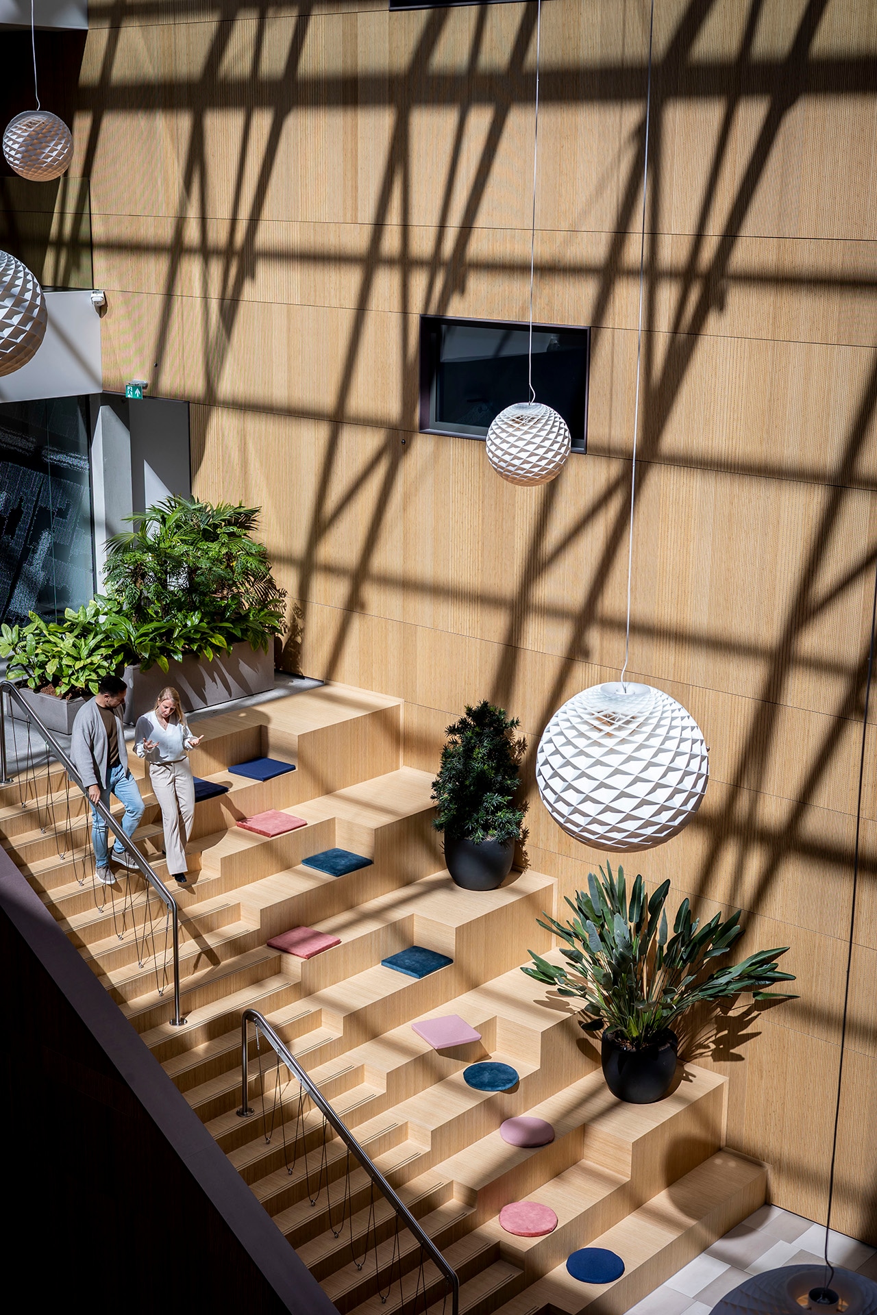 Moderne binnentrap met houten treden, kleurrijke zitkussens en planten in potten. Twee mensen praten met elkaar bij de trap. Grote bolvormige lampen hangen aan het plafond en werpen geometrische schaduwen op de houten muur.