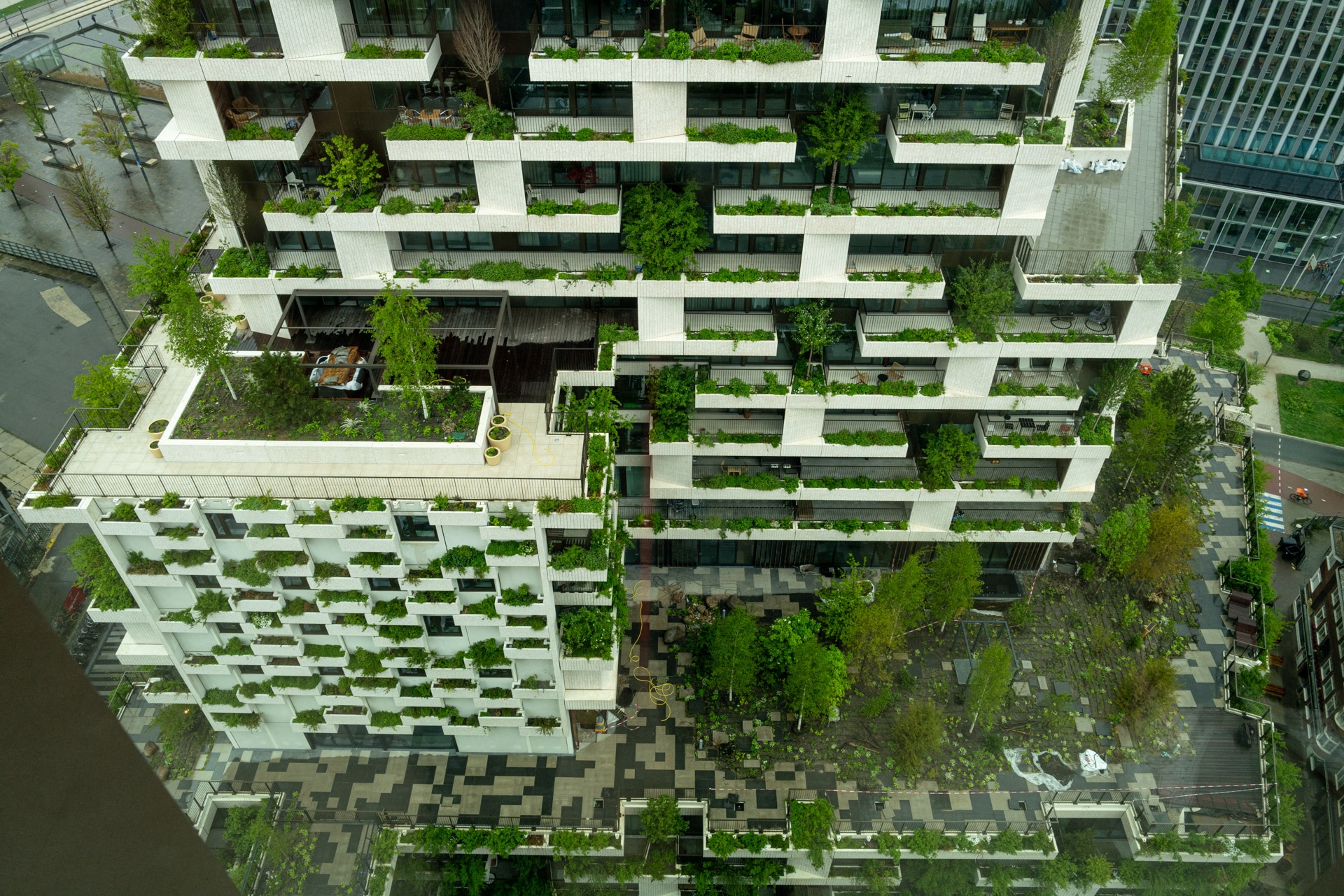 Luchtfoto van een modern gebouw met meerdere verdiepingen met talloze balkons en terrassen vol weelderige groene planten, omgeven door een binnenplaats met patroon en een stedelijk landschap.