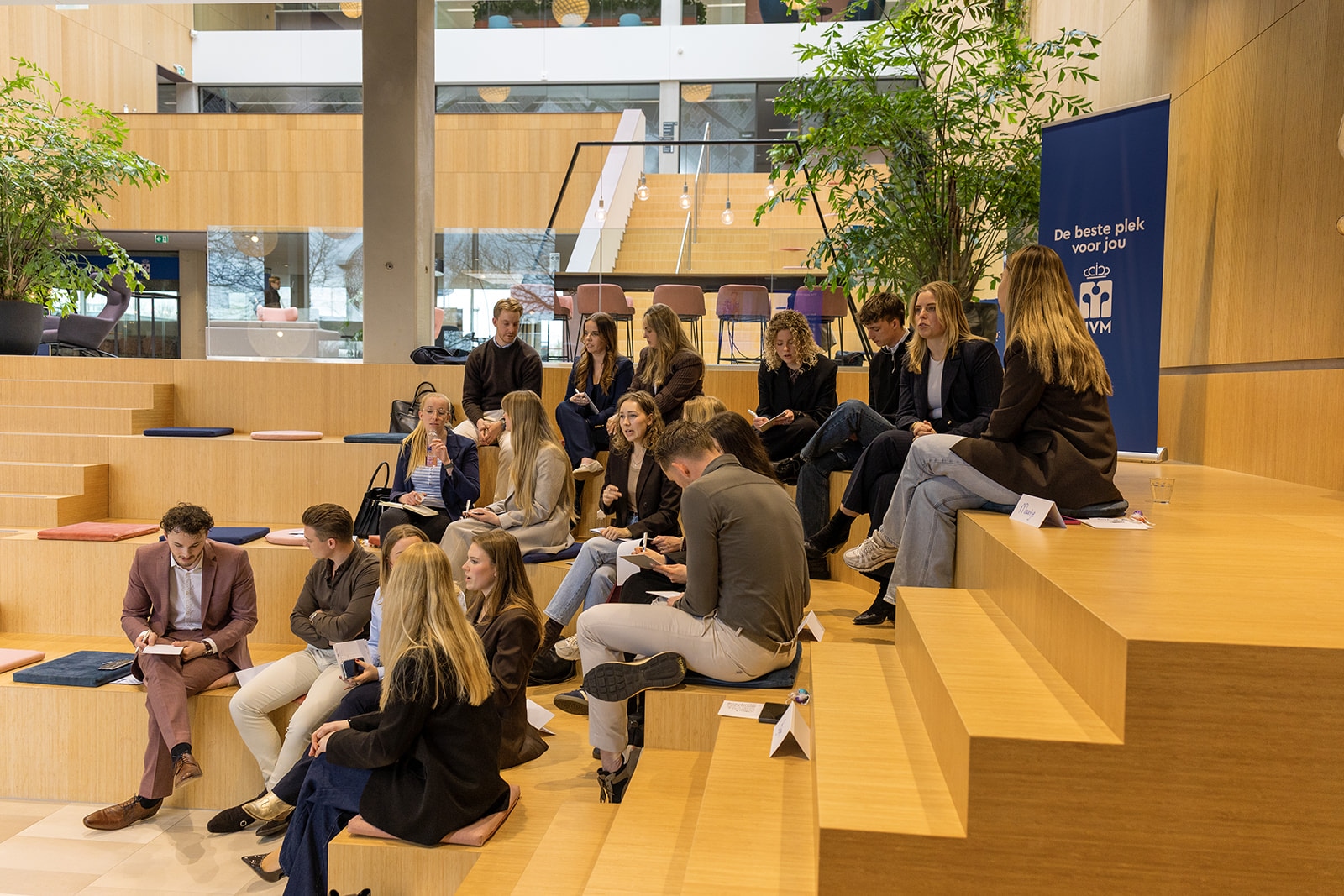 Een groep mensen zit en praat op moderne houten stoelen in een lichte, open binnenruimte met planten en grote ramen. Rechts staat een blauwe banner met Nederlandse tekst en een logo.