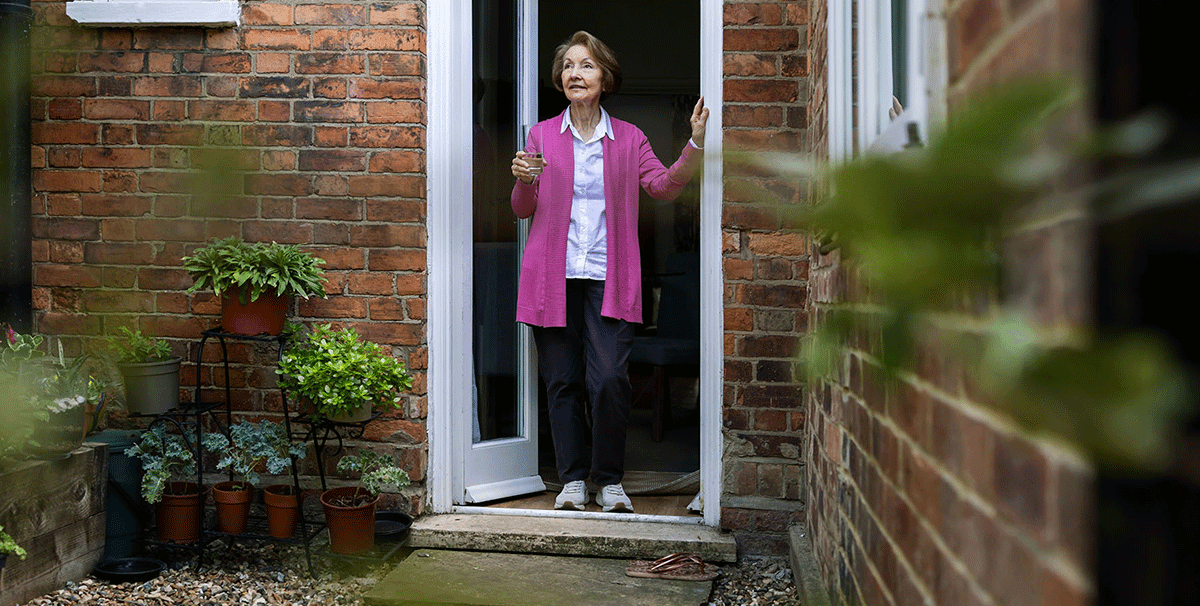 Een oudere vrouw staat in de deuropening van een bakstenen huis, houdt een drankje vast en kijkt naar buiten. Ze draagt een roze vest, een wit shirt en een donkere broek. Potplanten staan op een standaard in de buurt.