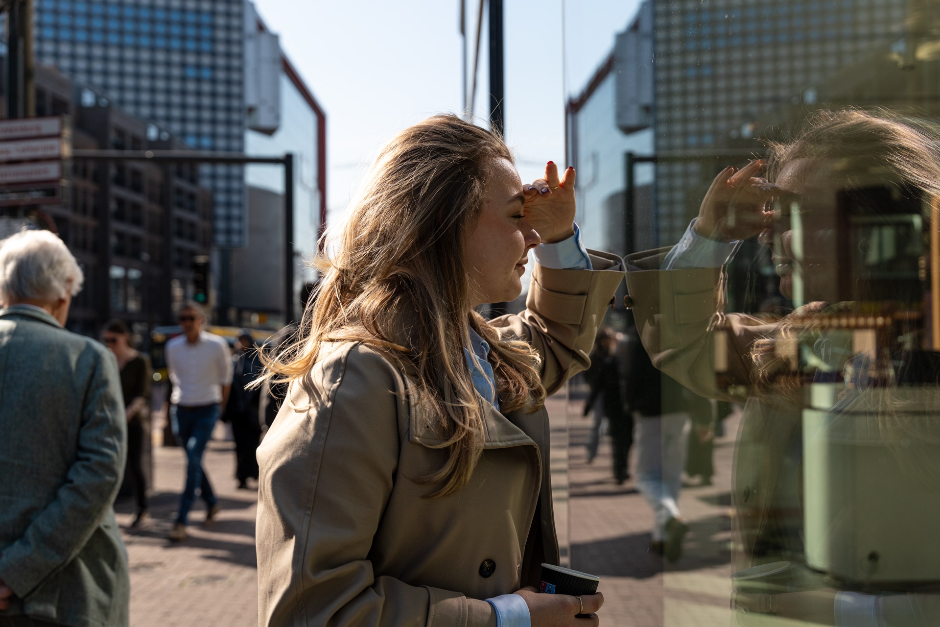 Een vrouw in een beige jas schermt haar ogen af van de zon terwijl ze door een glazen raam kijkt in een drukke stadsstraat, met lopende mensen en hoge gebouwen op de achtergrond.