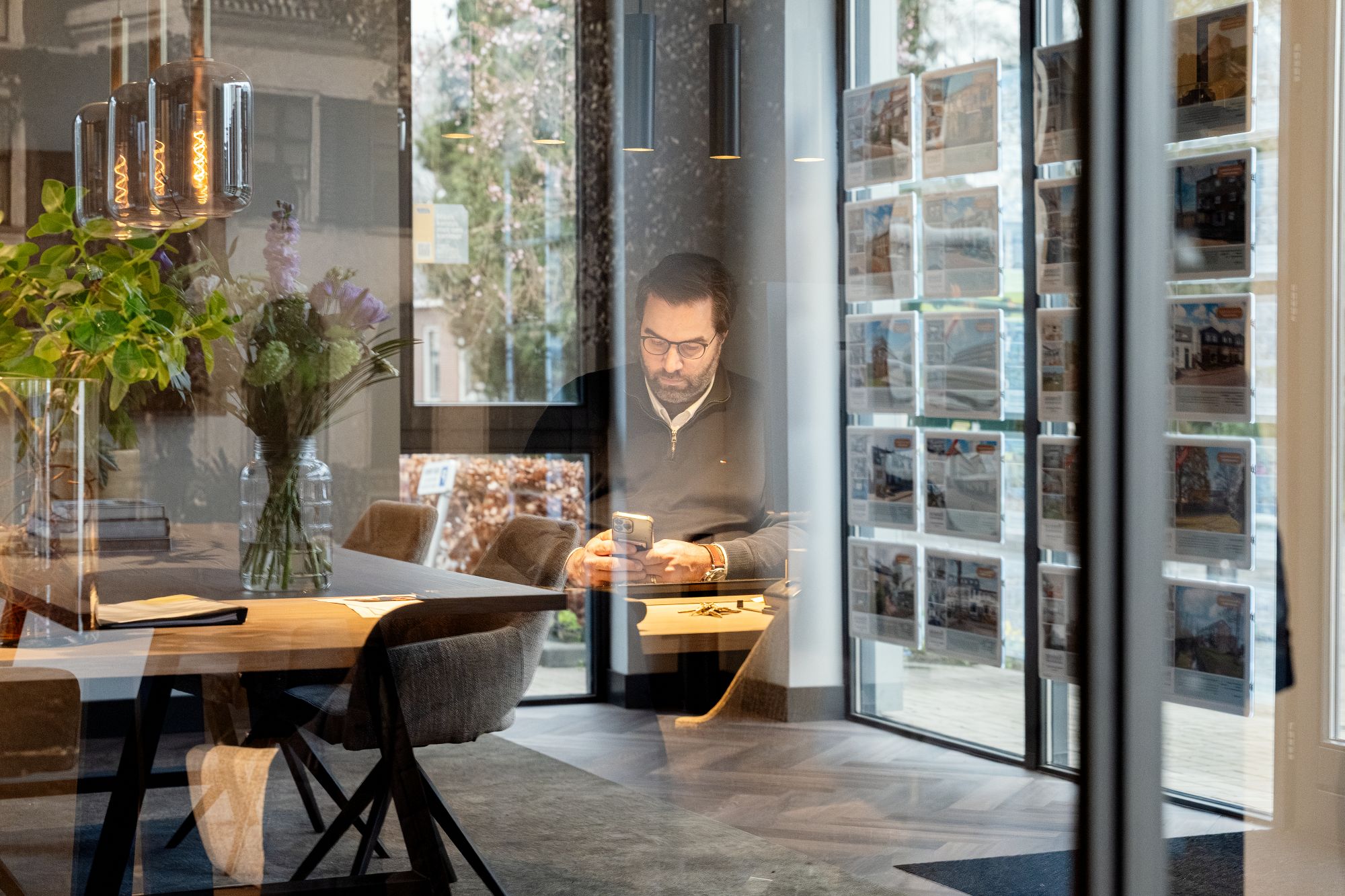 Een man zit alleen aan een tafel in een modern kantoor en kijkt naar zijn telefoon. Grote ramen weerspiegelen de buitenlucht en op de achtergrond zijn rekken met brochures of afbeeldingen te zien.