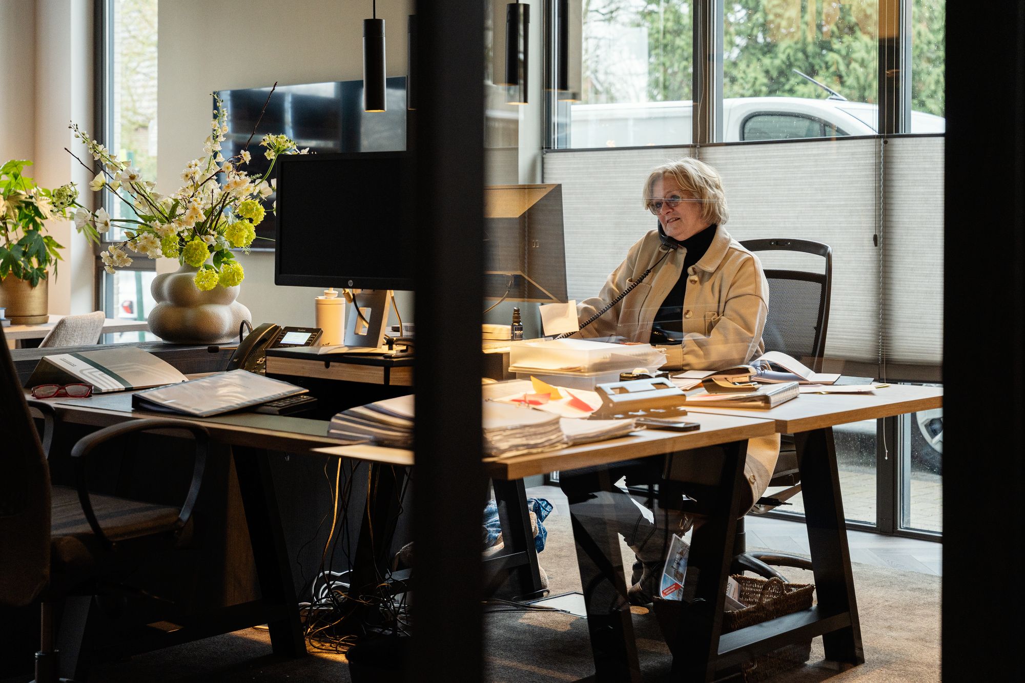Een vrouw zit aan een groot bureau in een modern kantoor, telefoneert en glimlacht. Papieren, boeken en kantoorbenodigdheden liggen verspreid over het bureau. Grote ramen zorgen voor natuurlijk licht en uitzicht naar buiten.