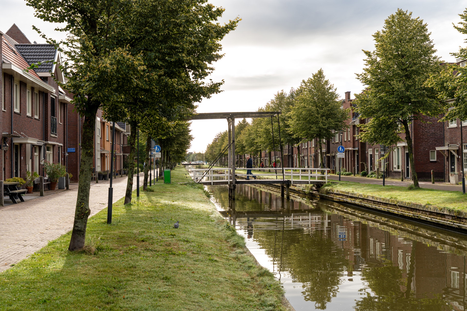 Een rustig kanaal omzoomd met bomen en bakstenen huizen aan beide zijden, met een kleine witte ophaalbrug in het midden onder een bewolkte hemel. Het water weerspiegelt de brug, de bomen en de omringende huizen.