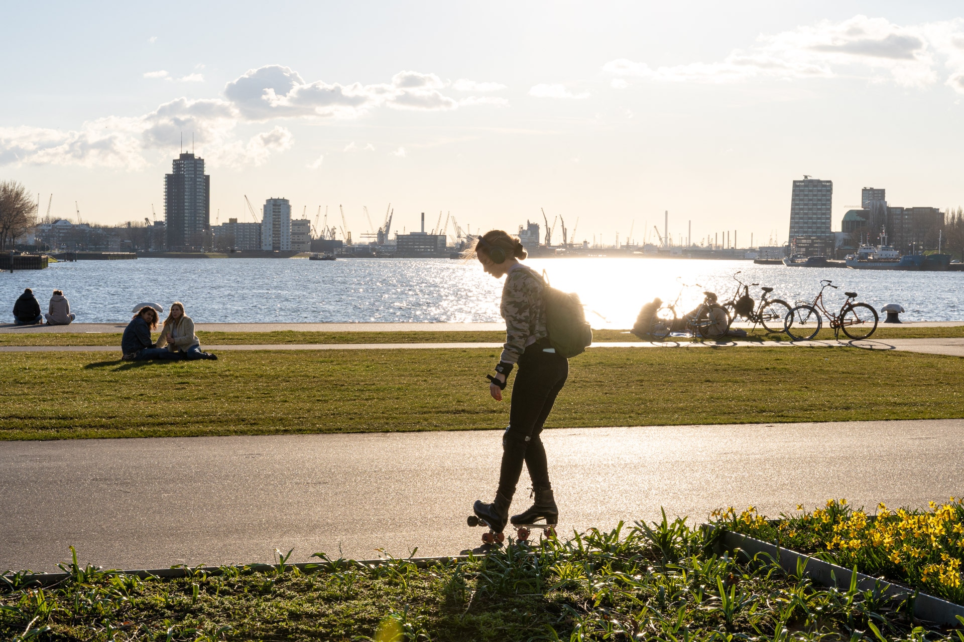 Een persoon rijdt op een skateboard langs een pad langs de rivier bij zonsondergang, met groepen mensen die op het gras zitten en fietsen die vlakbij geparkeerd staan; stadsgebouwen en kranen zijn zichtbaar aan de overkant van het water.