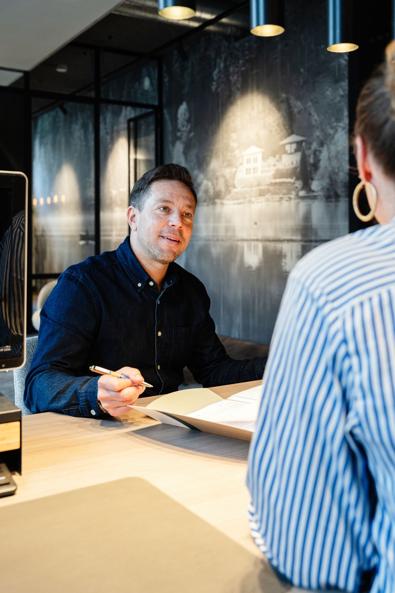 Een man in een donker shirt zit aan een bureau, houdt een pen vast en glimlacht terwijl hij praat met een vrouw in een gestreept shirt. Op het bureau liggen papieren en op de achtergrond een grote muurschildering van een huis aan een meer.
