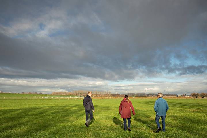 Drie mensen lopen over een breed, groen grasveld onder een bewolkte hemel, met in de verte bomen en landbouwgrond zichtbaar aan de horizon.
