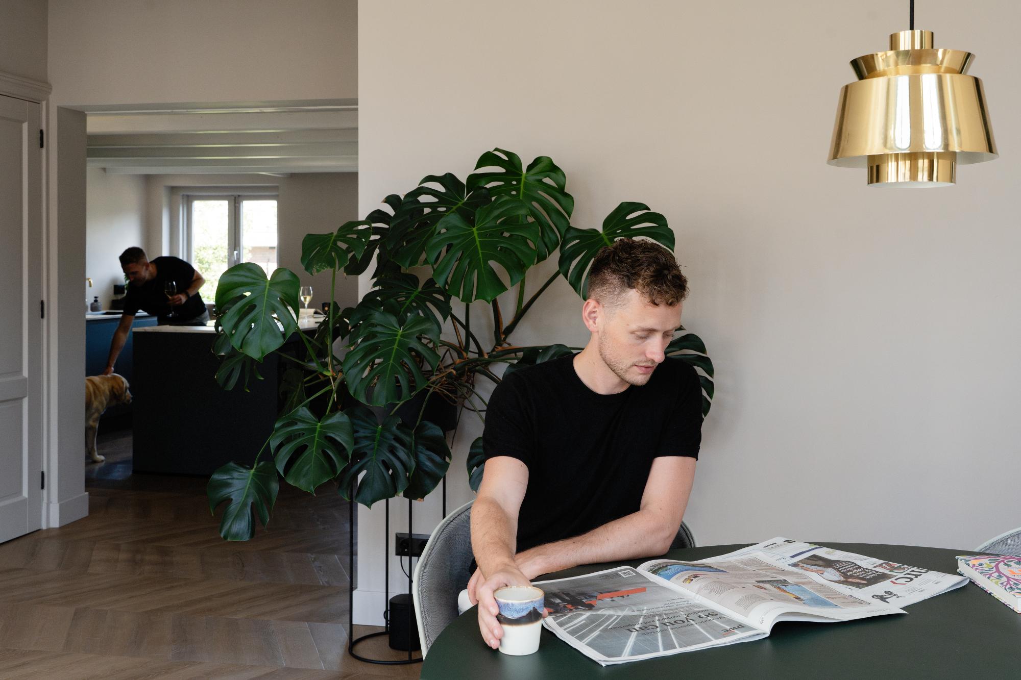 Een man in een zwart overhemd zit aan een tafel een tijdschrift te lezen en een kopje vast te houden. Achter hem zijn een grote bladplant en een gouden lamp zichtbaar, met op de achtergrond twee mensen en een hond bij de keuken.