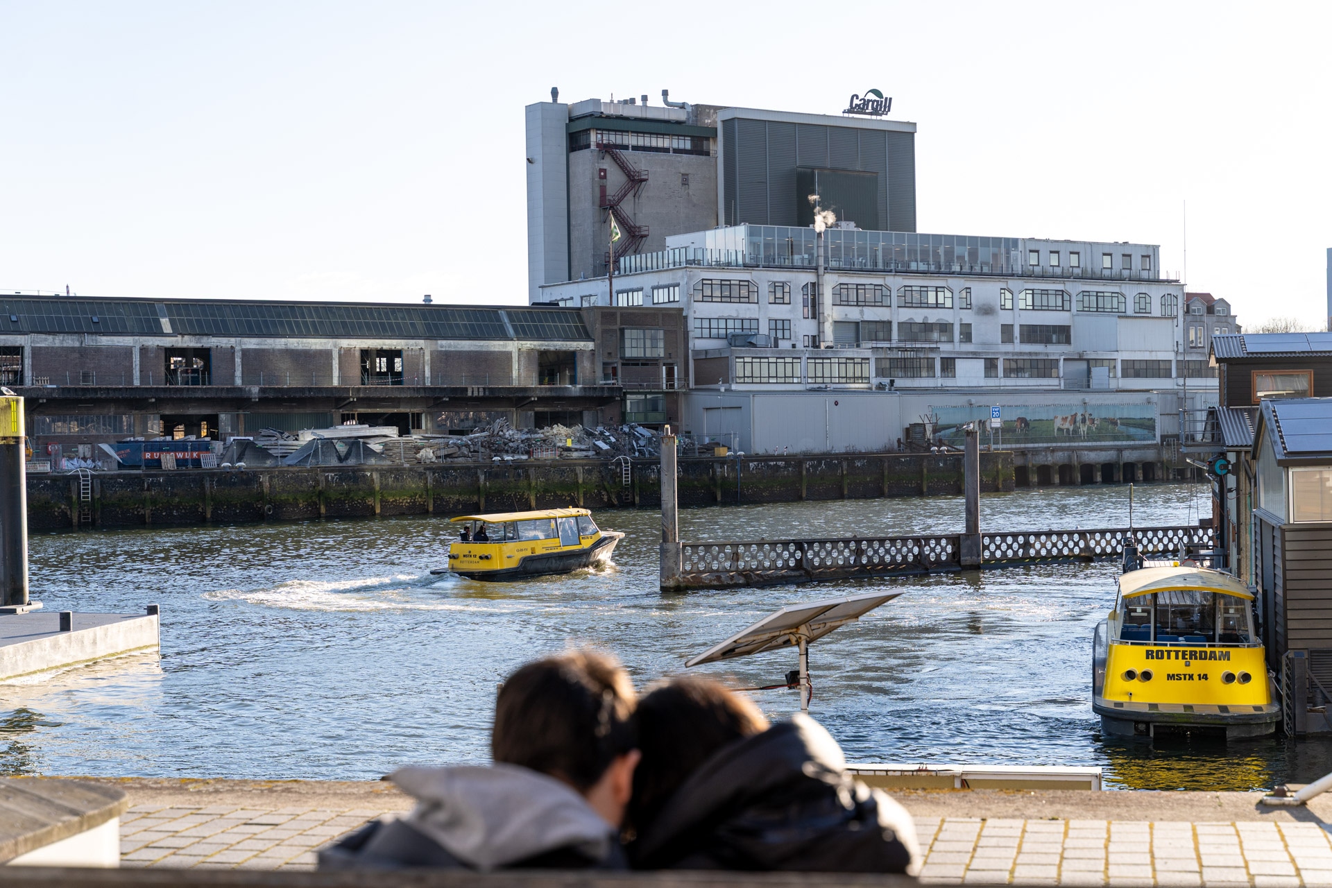 Twee mensen zitten aan het water in Rotterdam en kijken naar gele watertaxi's die over de rivier rijden. Industriële gebouwen en een pier zijn zichtbaar op de achtergrond onder een heldere hemel.